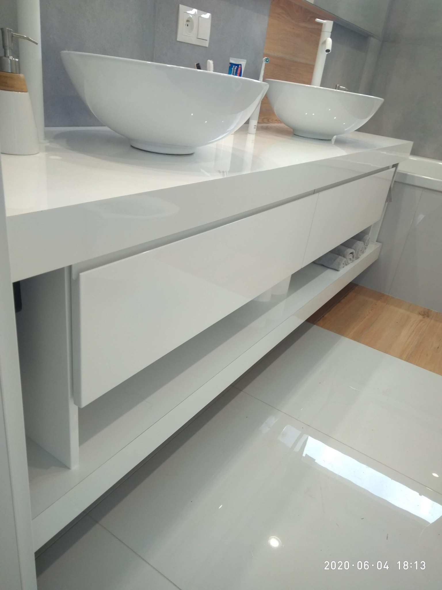 Nowoczesna łazienka z dwiema białymi, okrągłymi umywalkami nablatowymi na białej szafce z szufladami i otwartą półką z ręcznikami, minimalistyczny design, białe płytki podłogowe.