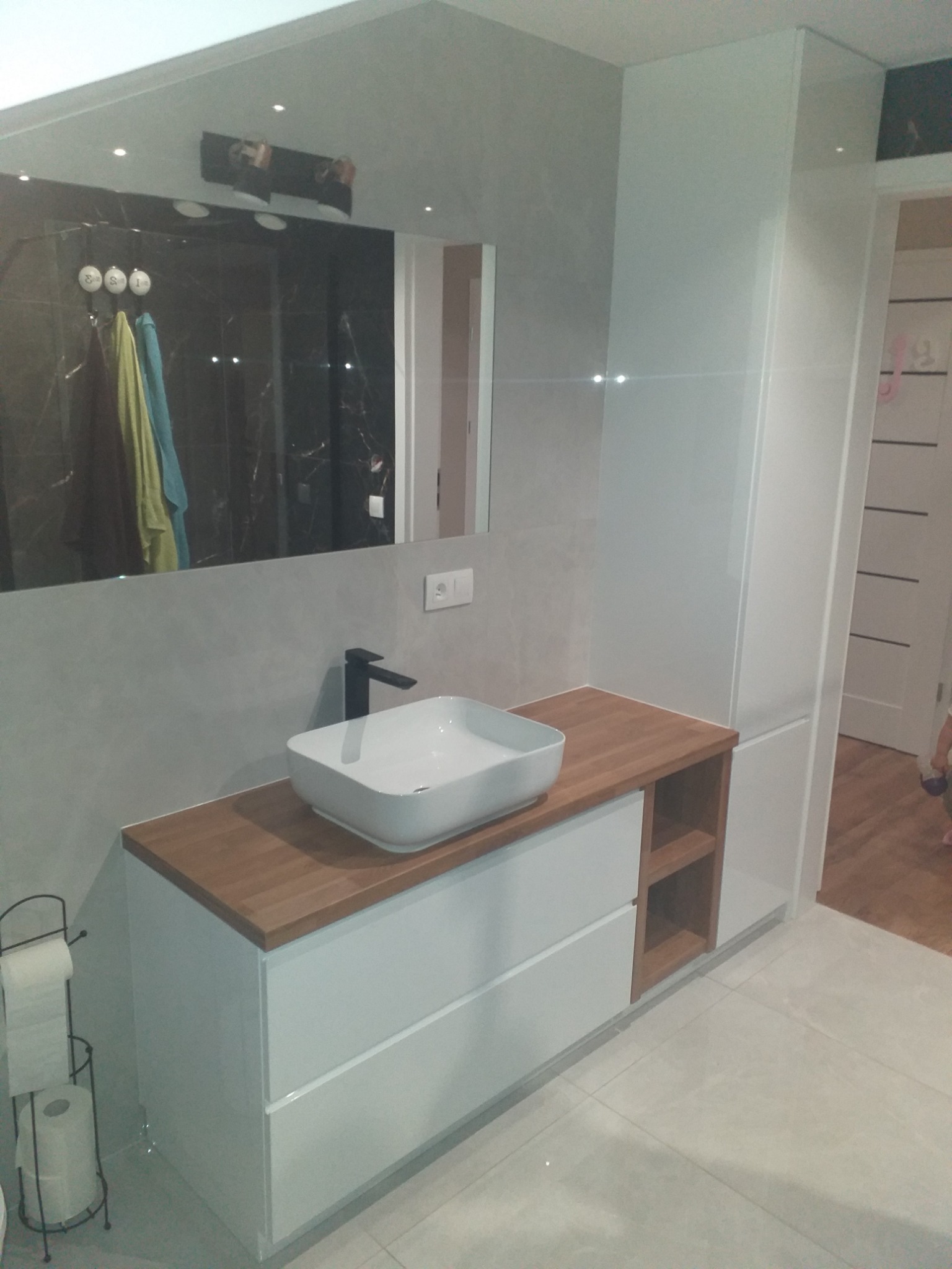 Nowoczesna łazienka z białą szafką pod umywalkę z drewnianym blatem, czarną armaturą i dużym lustrem odbijającym kabinę prysznicową z czarnymi płytkami.