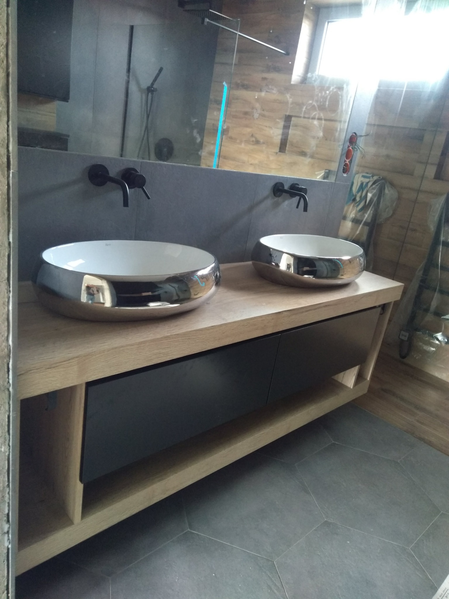 Dwie okrągłe umywalki z chromowanymi brzegami stojące na drewnianym blacie z czarnymi szafkami w nowoczesnej łazience z szarymi płytkami i czarną armaturą.