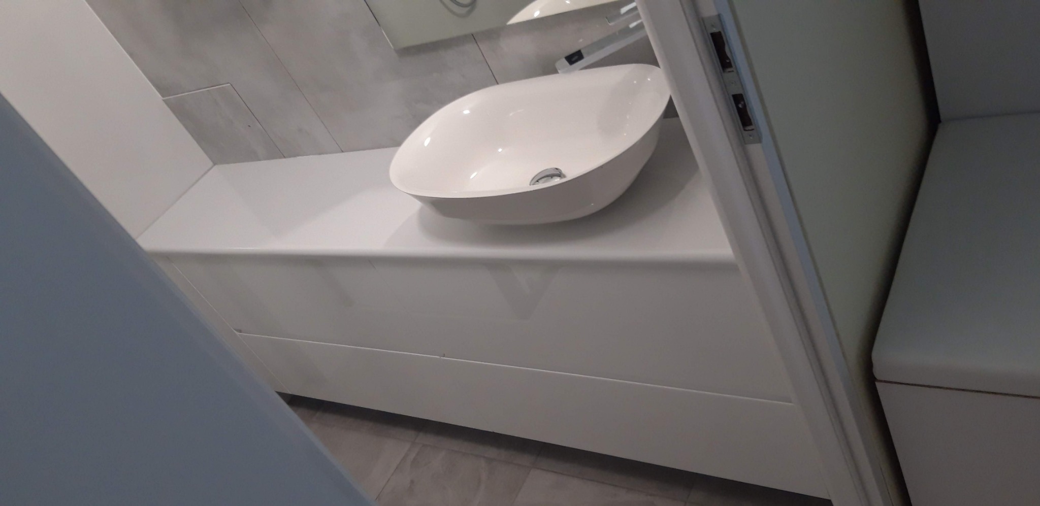 Biała, owalna umywalka nablatowa zamontowana na białym, minimalistycznym blacie w łazience, widoczna zza uchylonych drzwi.