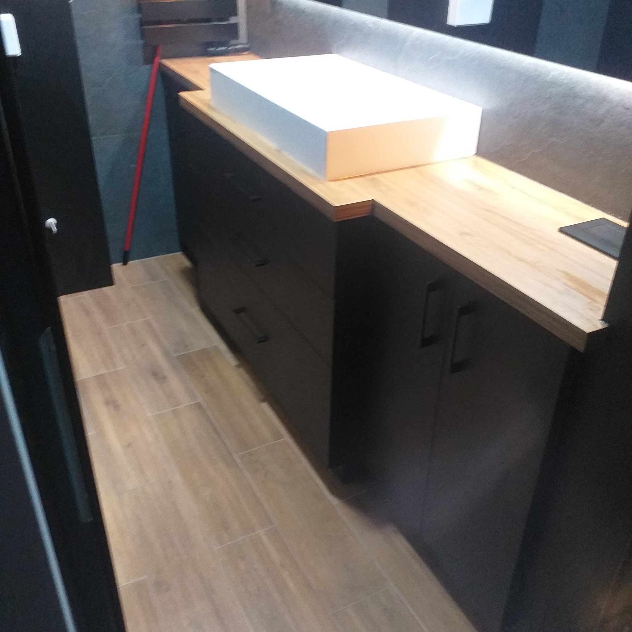 Nowoczesna łazienka z szafką pod umywalkę w kolorze ciemnego grafitu, blatem z jasnego drewna i prostokątną białą umywalką nablatową.