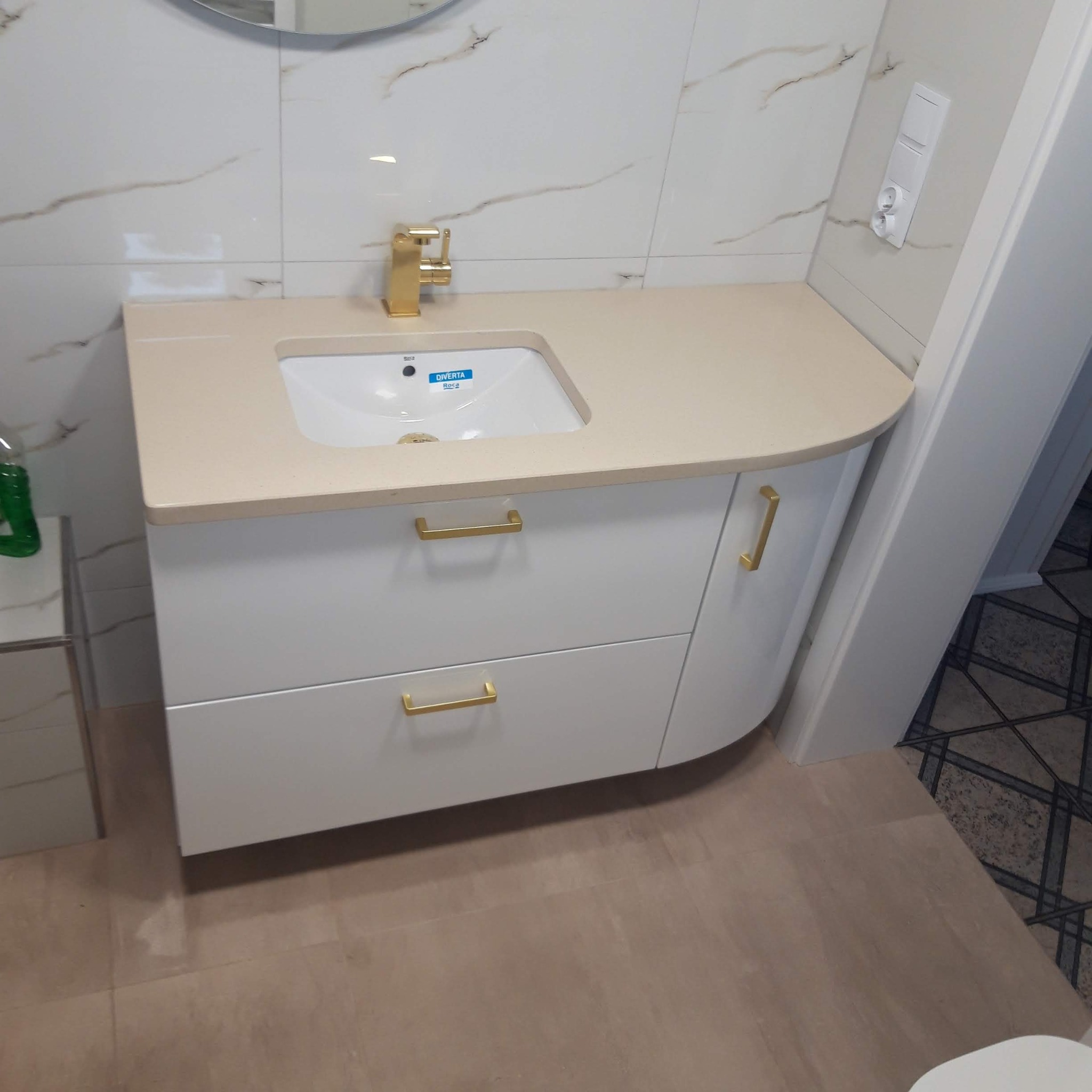 Nowoczesna łazienka z białą szafką pod umywalkę z beżowym blatem i złotymi uchwytami, marmurowe płytki na ścianie, złota bateria.