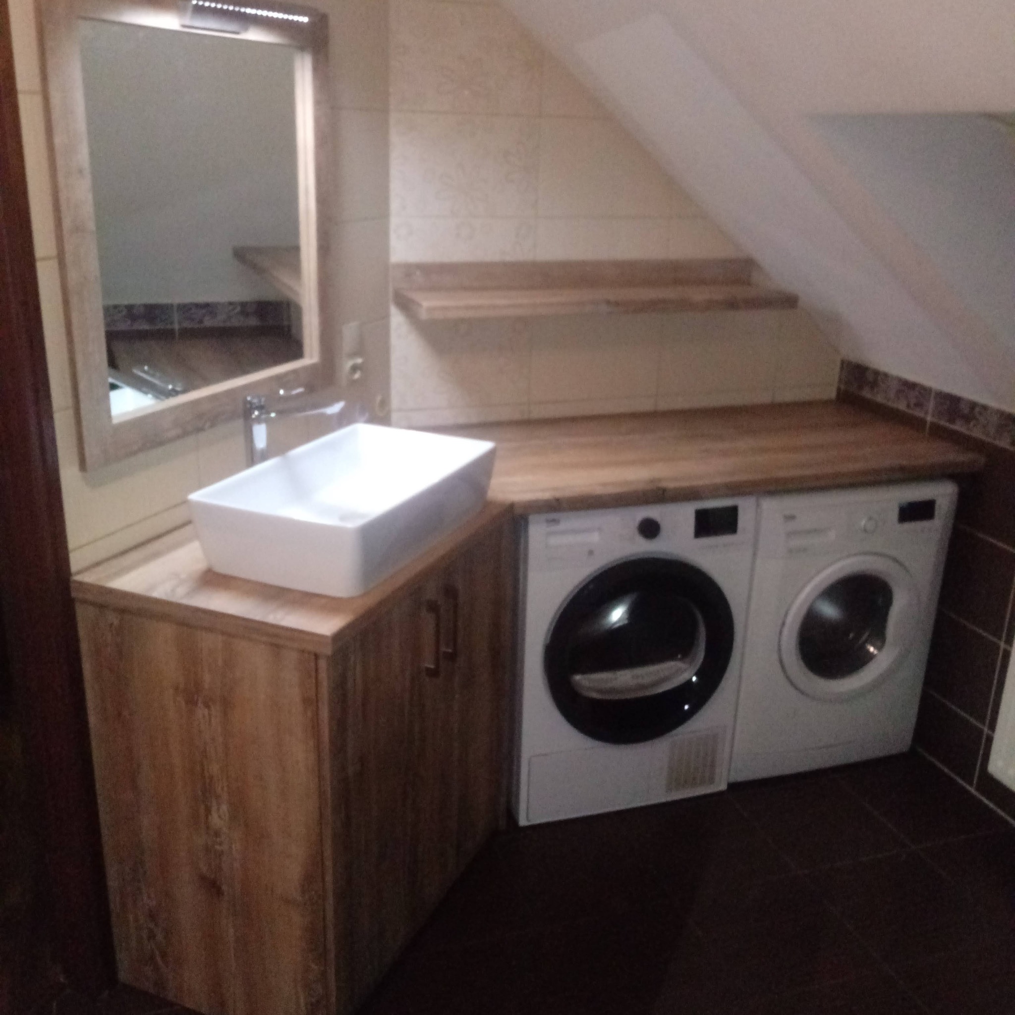 Łazienka na poddaszu z drewnianym blatem, na którym stoi prostokątna umywalka i dwie pralki; lustro z oświetleniem nad umywalką.