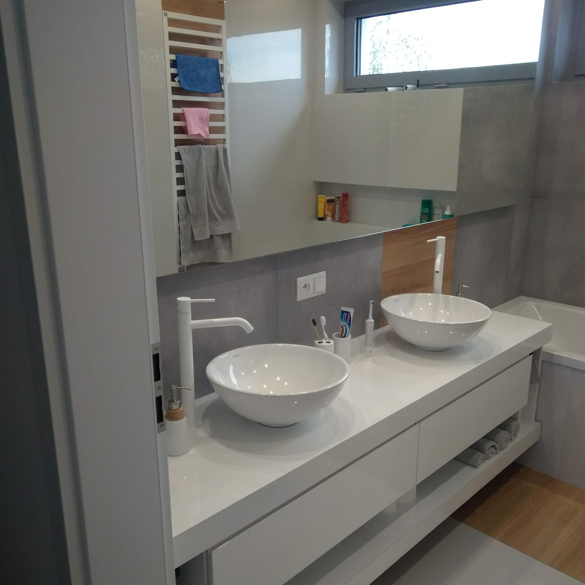 Nowoczesna łazienka z dwoma białymi umywalkami nablatowymi na białym blacie z szufladami, biała armatura, lustro z grzejnikiem drabinkowym i ręcznikami w tle.