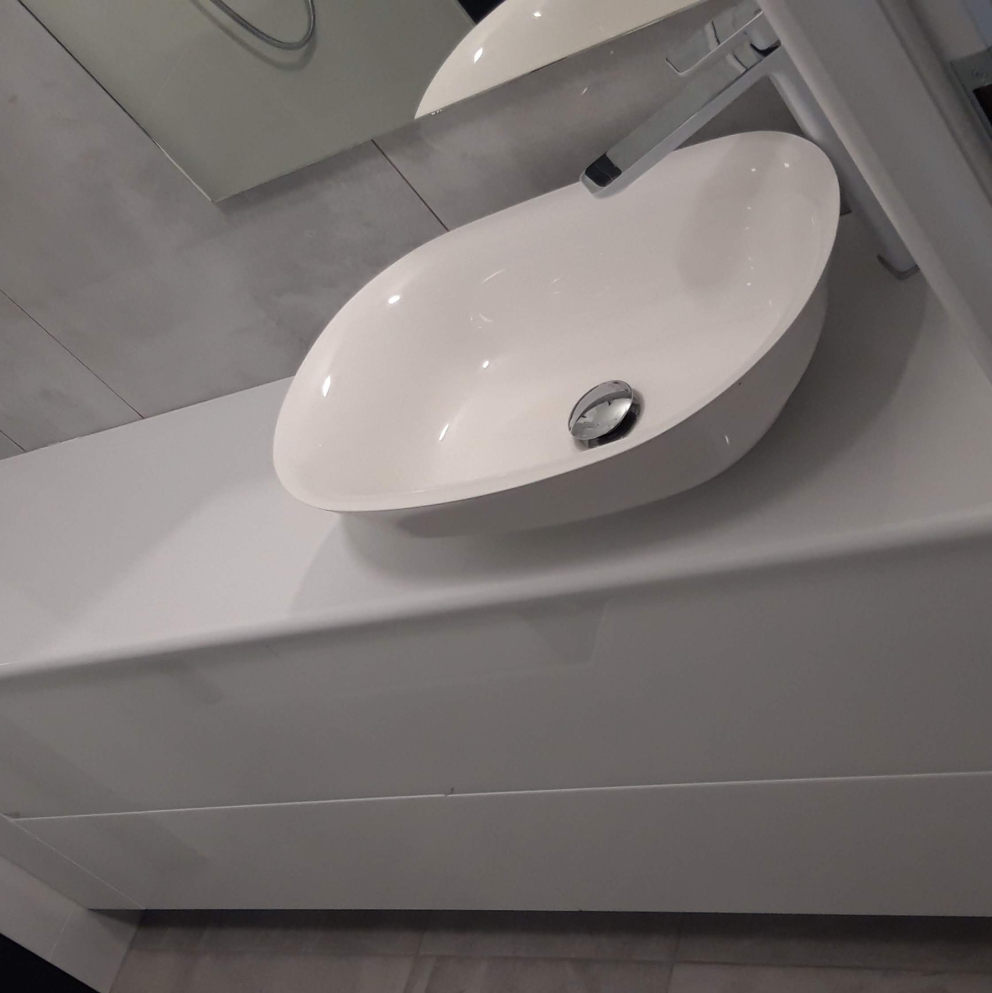 Nowoczesna, owalna umywalka nablatowa z chromowaną baterią, zamontowana na białym, minimalistycznym blacie z szufladami, na tle szarej ściany wykończonej płytkami imitującymi beton.