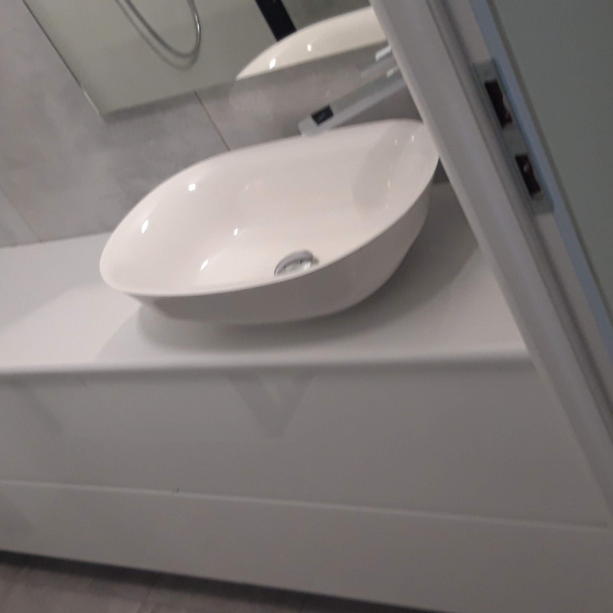 Nowoczesna, biała umywalka nablatowa o owalnym kształcie z chromowanym odpływem, zamontowana na białym blacie w minimalistycznej łazience z szarą płytką na ścianie i częściowo widoczną kabiną...