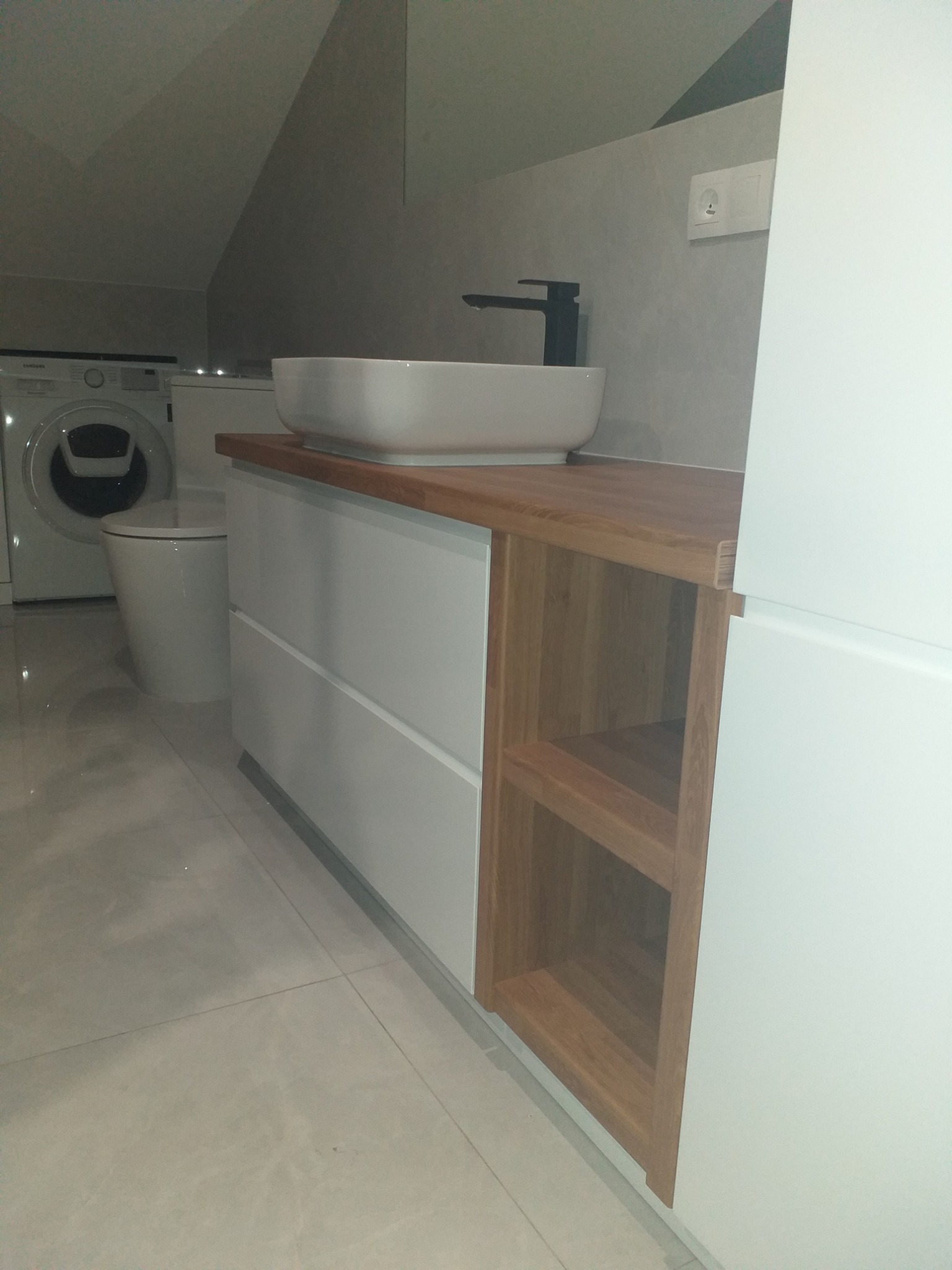 Biała umywalka nablatowa na szafce z szufladami i drewnianym blatem w łazience na poddaszu, obok pralka i toaleta.