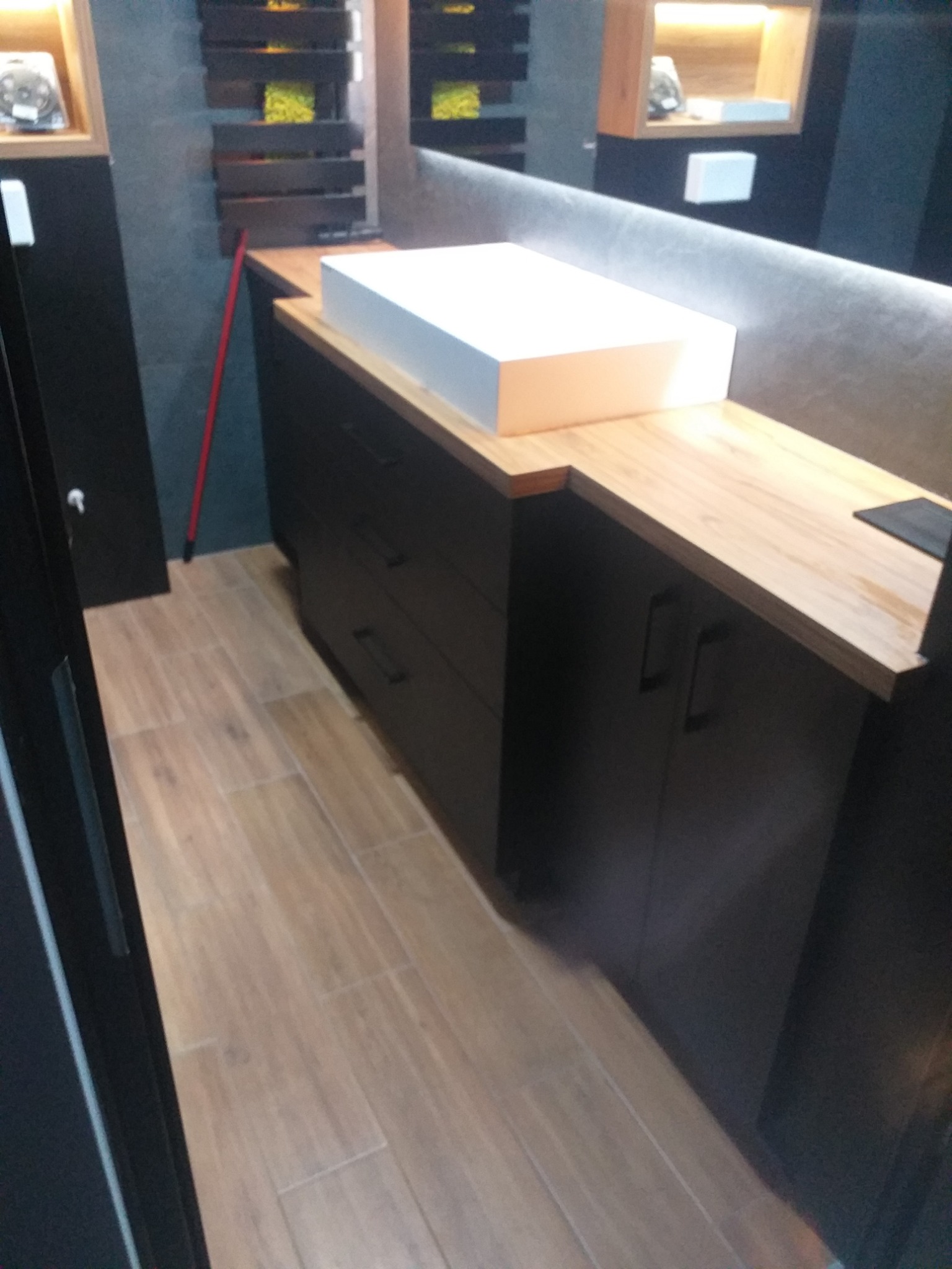 Ciemne szafki łazienkowe z drewnianym blatem i białą umywalką nablatową, widok z boku, na podłodze płytki imitujące drewno, w tle dekoracyjne półki z oświetleniem.