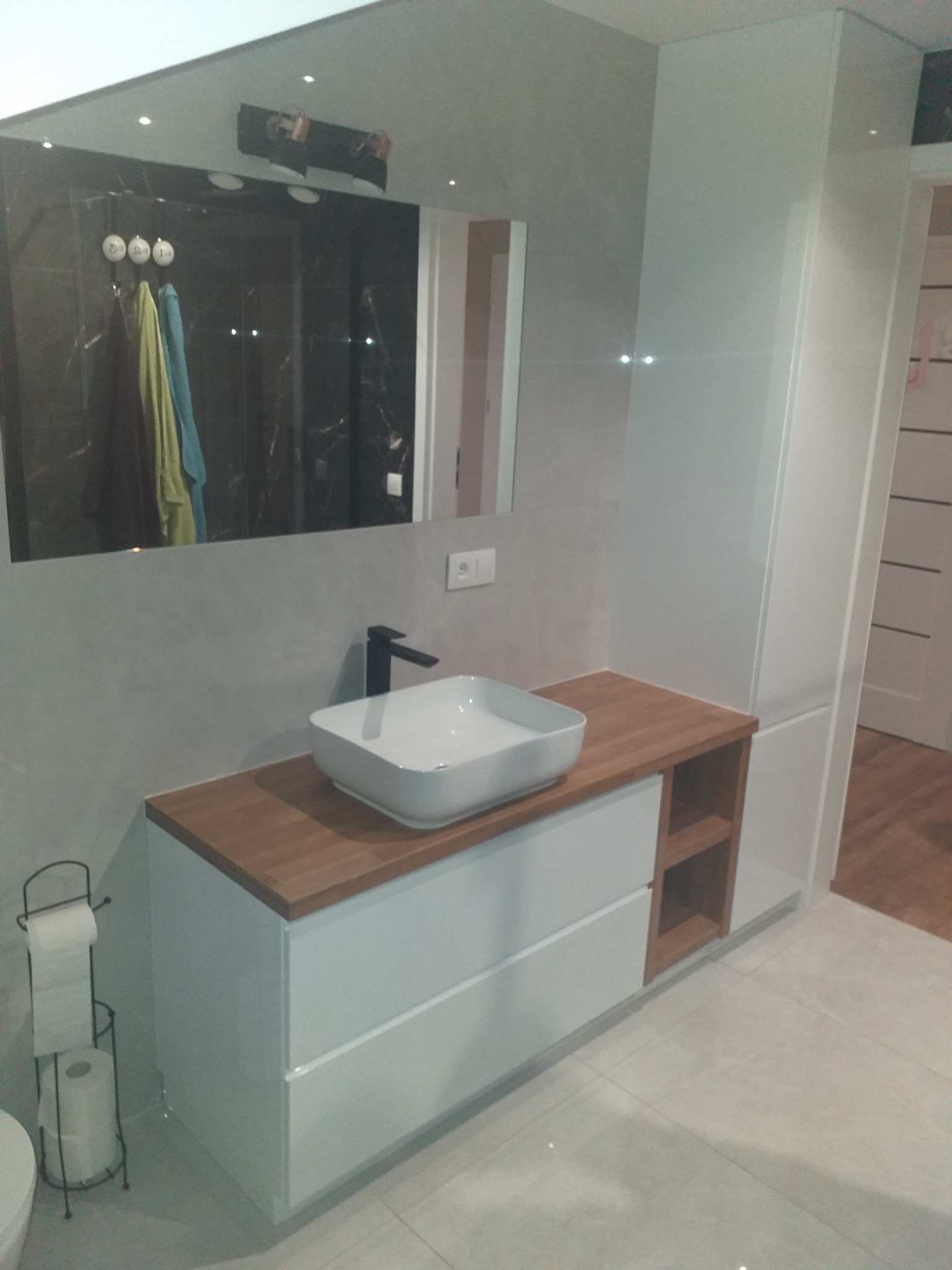 Nowoczesna łazienka z białą szafką pod umywalkę z drewnianym blatem, minimalistyczna armatura, duże lustro odbijające czarną ścianę prysznica i akcesoria łazienkowe.