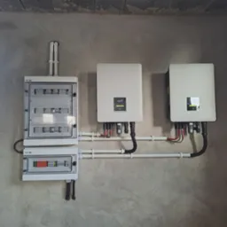 Dwa białe inwertery fotowoltaiczne FOX ESS zamontowane na surowej, betonowej ścianie obok dwóch skrzynek elektrycznych, z widocznymi przewodami i rurami instalacyjnymi.