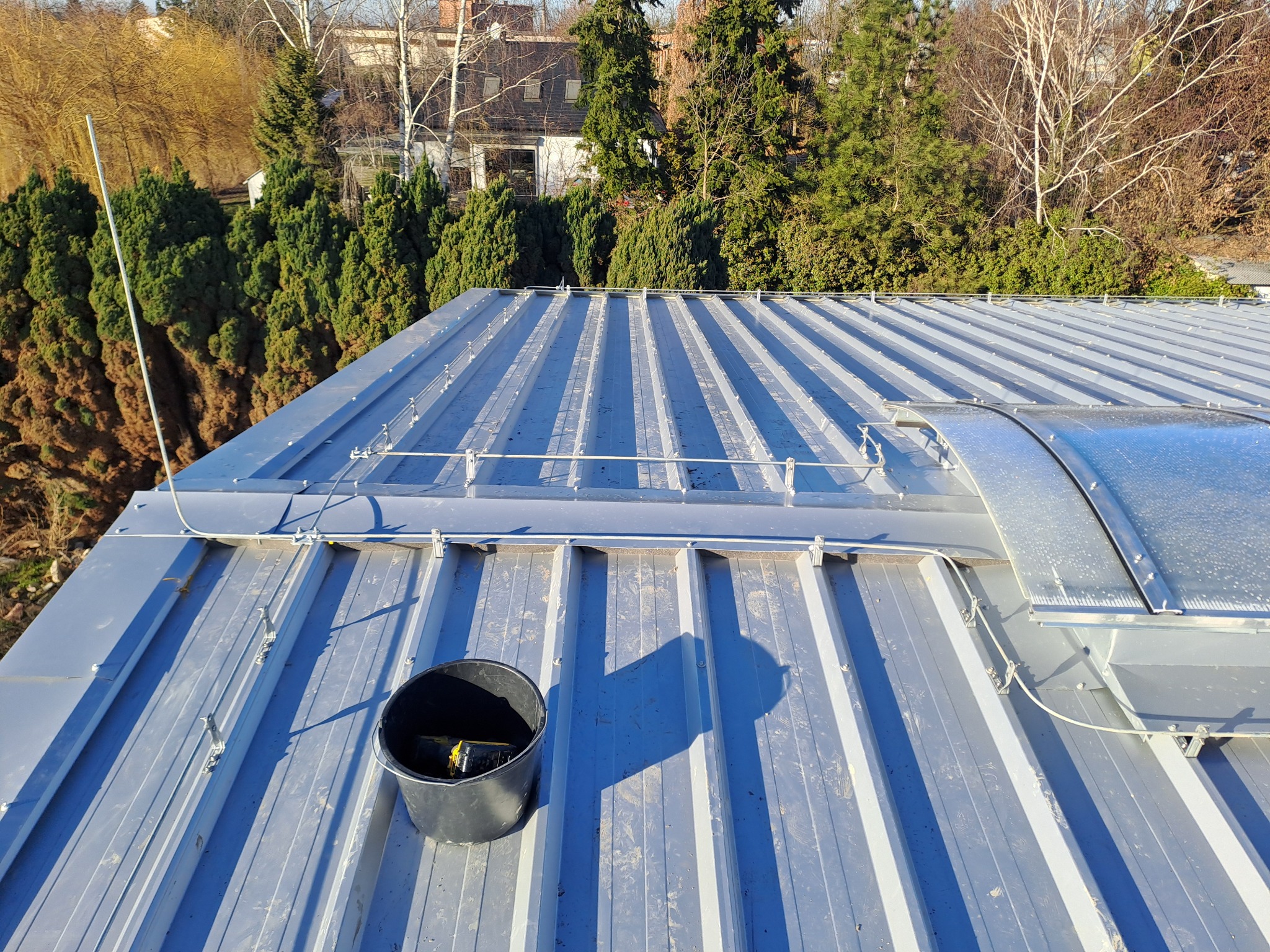 Blaszany dach z zamontowanymi uchwytami, przewodami i świetlikiem dachowym; czarne wiadro z narzędziami na pierwszym planie, drzewa w tle.