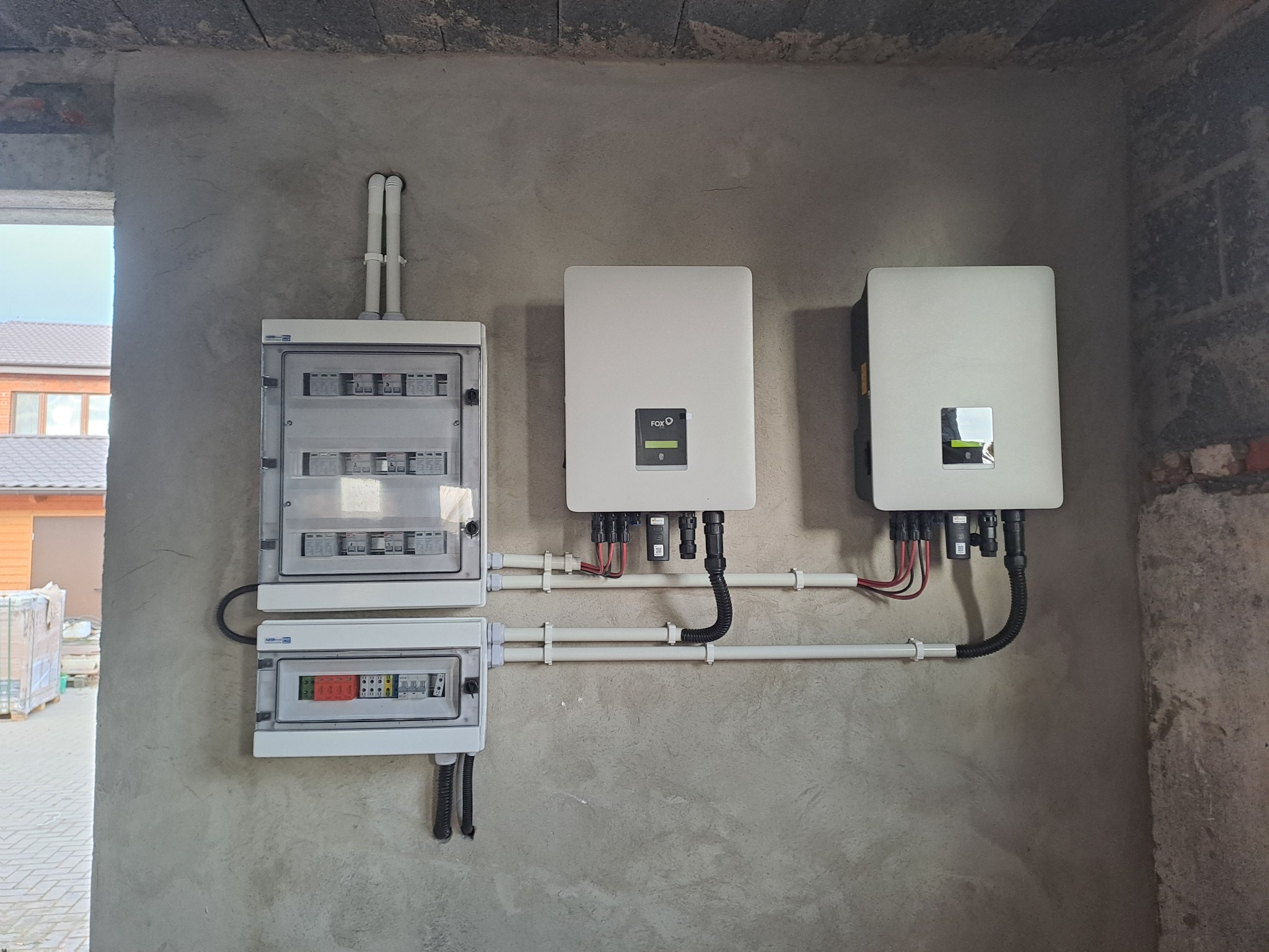 Dwa białe inwertery fotowoltaiczne FOX ESS zamontowane na surowej, betonowej ścianie obok dwóch skrzynek elektrycznych, z widocznymi przewodami i rurami instalacyjnymi.