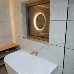 PICCOLI LAVORI - Biała wanna wolnostojąca z złotą armaturą na tle marmurowych płytek w łazience. W tle okno z okrągłym, podświetlanym lustrem.