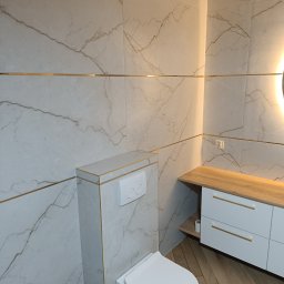 PICCOLI LAVORI - Elegancka łazienka z płytkami imitującymi marmur i złotymi akcentami, wisząca toaleta, szafka z drewnianym blatem i oświetleniem LED. Parkiet ułożony w jodełkę.