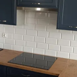 Białe, prostokątne płytki na ścianie nad blatem i płytą indukcyjną. Granatowe szafki z uchwytami. Metalowy okap. Gniazdko elektryczne po lewej.