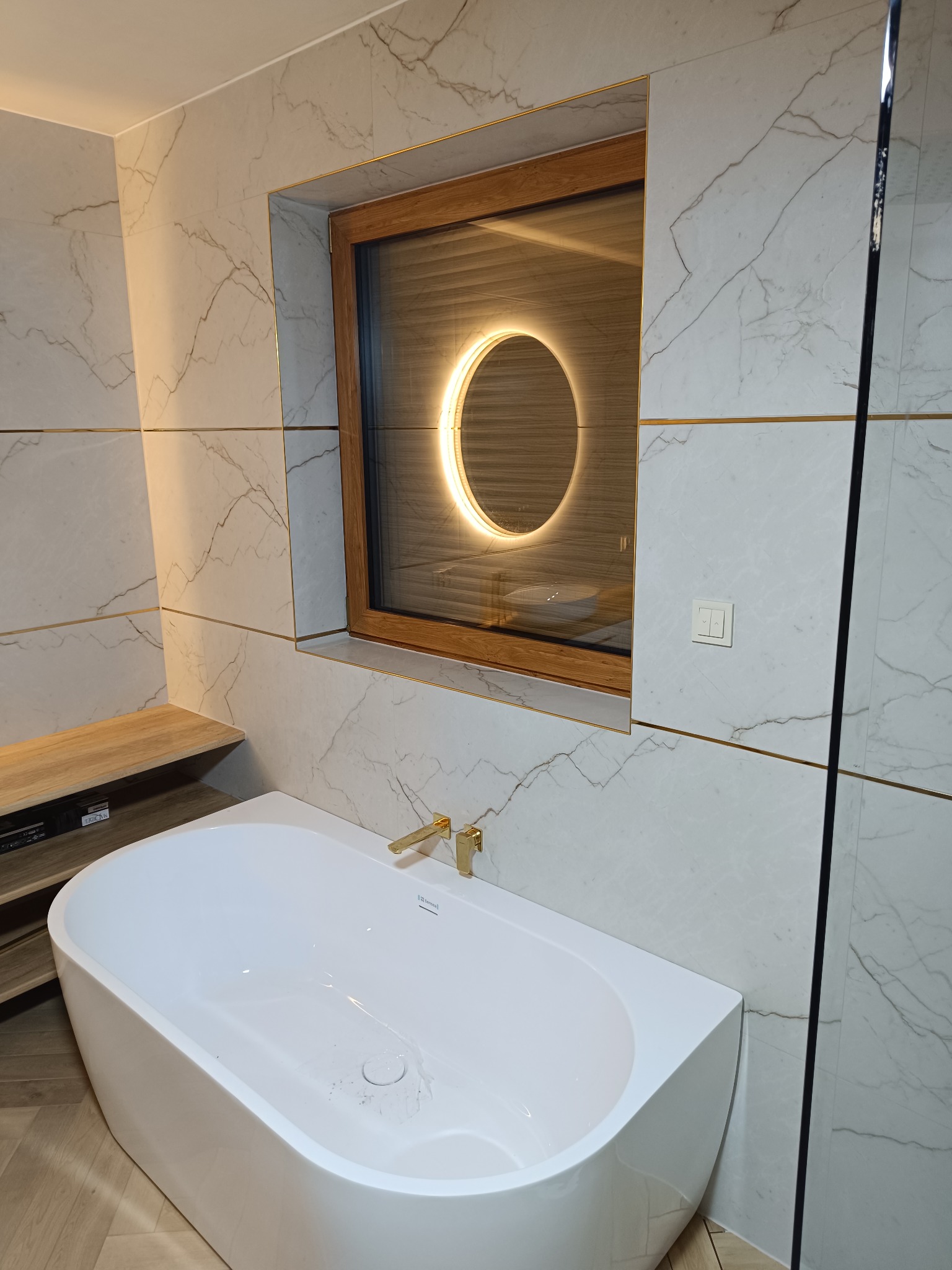 Biała wanna wolnostojąca z złotą armaturą na tle marmurowych płytek w łazience. W tle okno z okrągłym, podświetlanym lustrem.