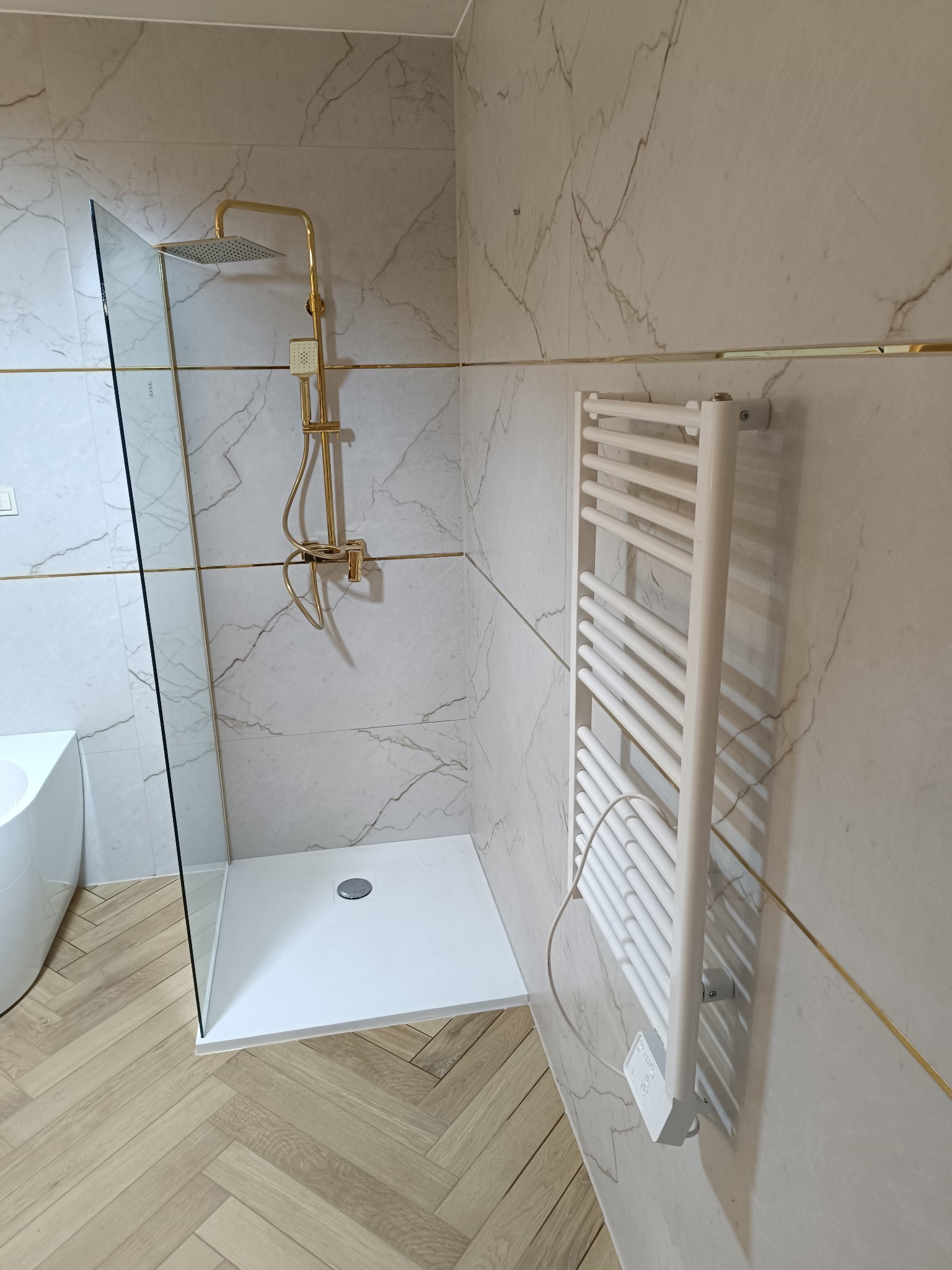 Nowoczesna łazienka z kabiną typu walk-in, złotą armaturą i grzejnikiem drabinkowym na marmurowej ścianie. Parkiet ułożony w jodełkę.