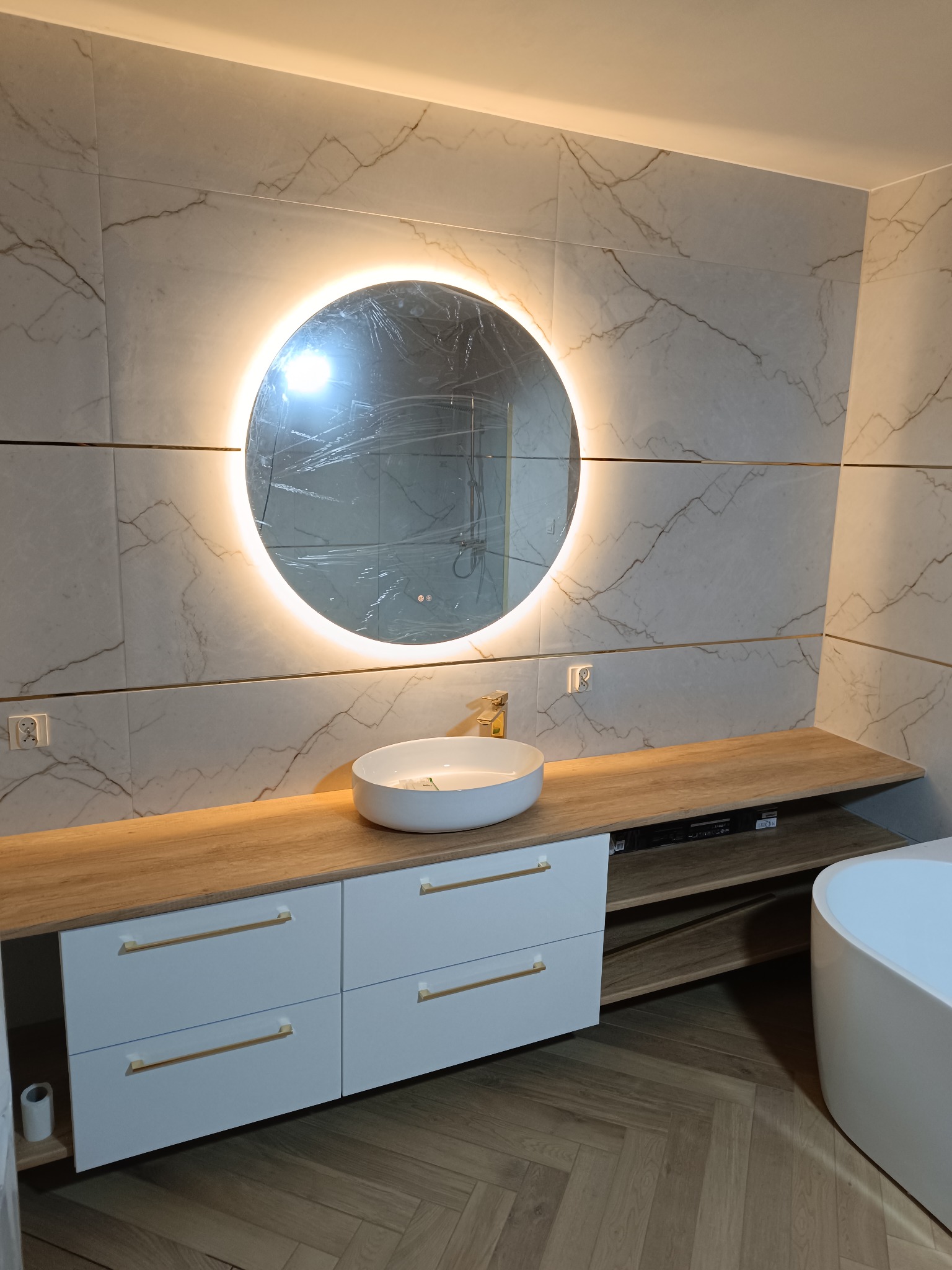 Nowoczesna łazienka: marmurowe płytki, okrągłe lustro z podświetleniem, biała umywalka na szafce z drewnianym blatem i złotymi uchwytami, parkiet w jodełkę.