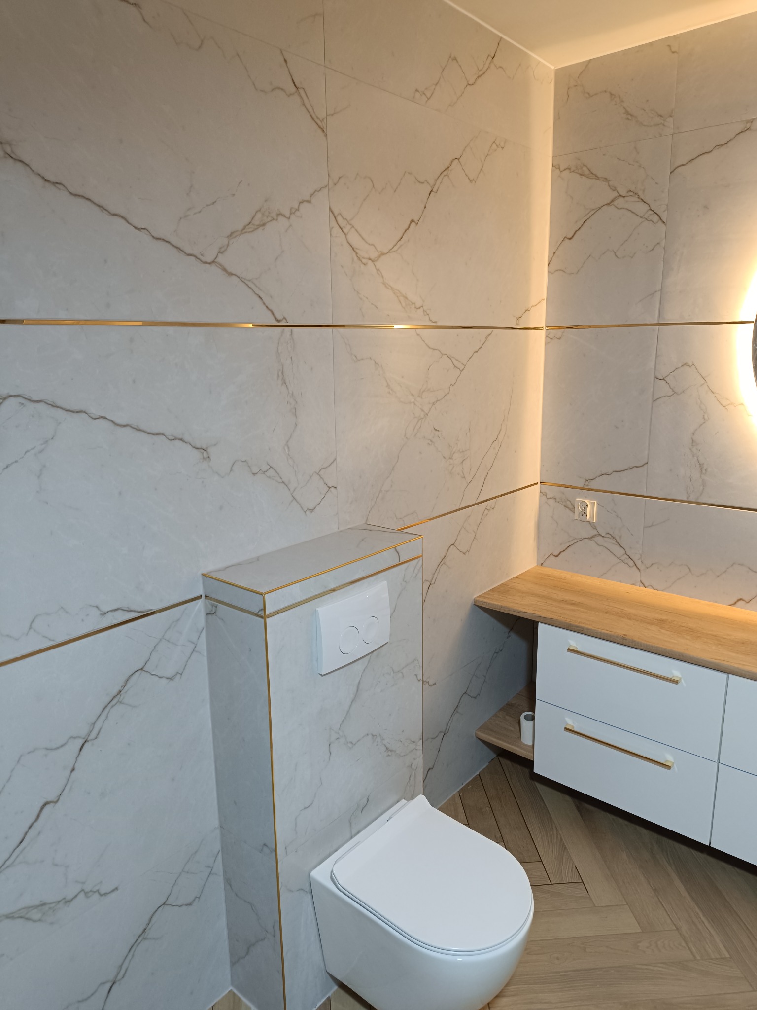 Elegancka łazienka z płytkami imitującymi marmur i złotymi akcentami, wisząca toaleta, szafka z drewnianym blatem i oświetleniem LED. Parkiet ułożony w jodełkę.