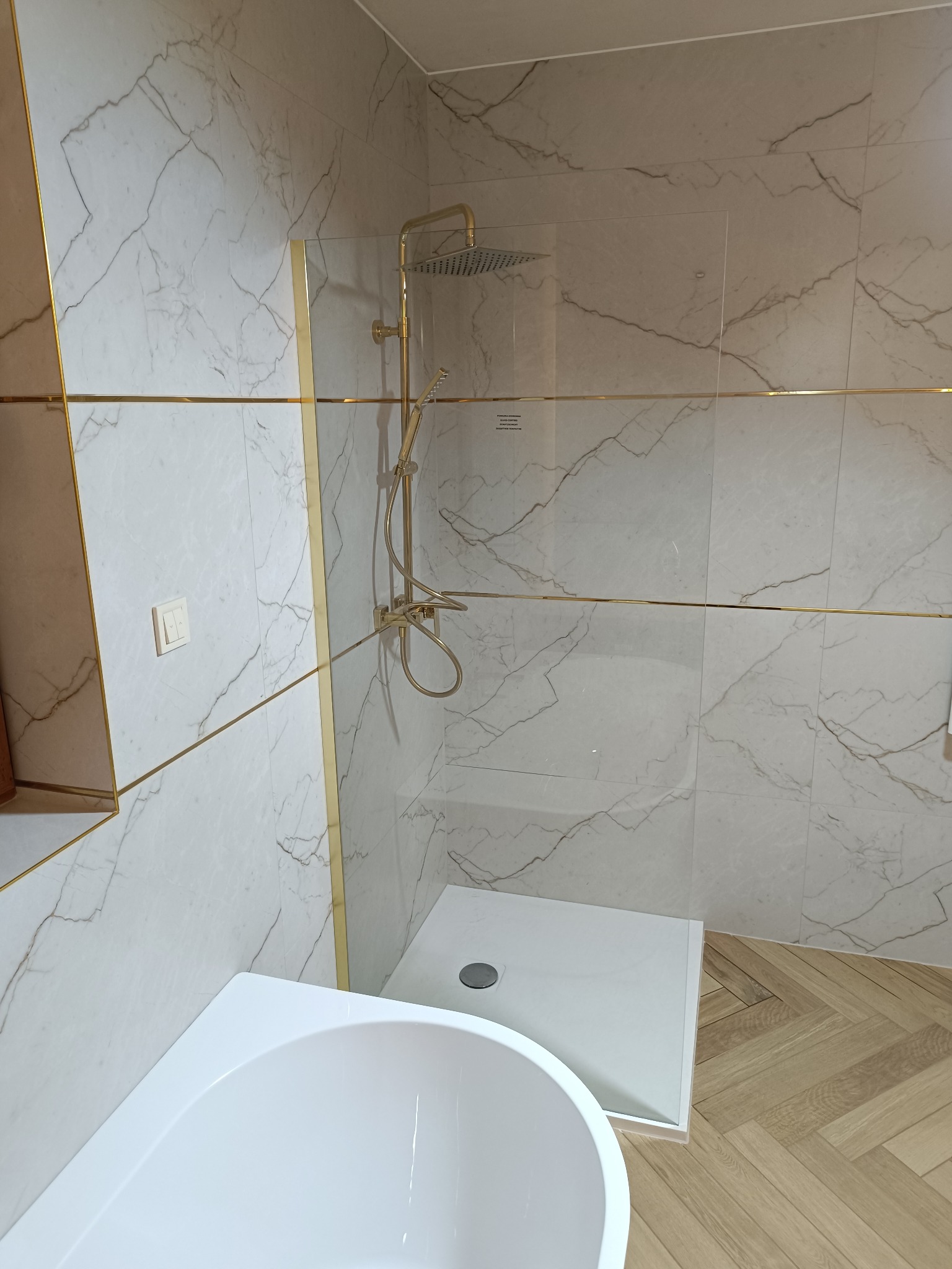 Biała, marmurkowa łazienka z kabiną typu walk-in, złotymi akcentami i drewnianą podłogą w jodełkę. Widoczna część wanny i deszczownica.