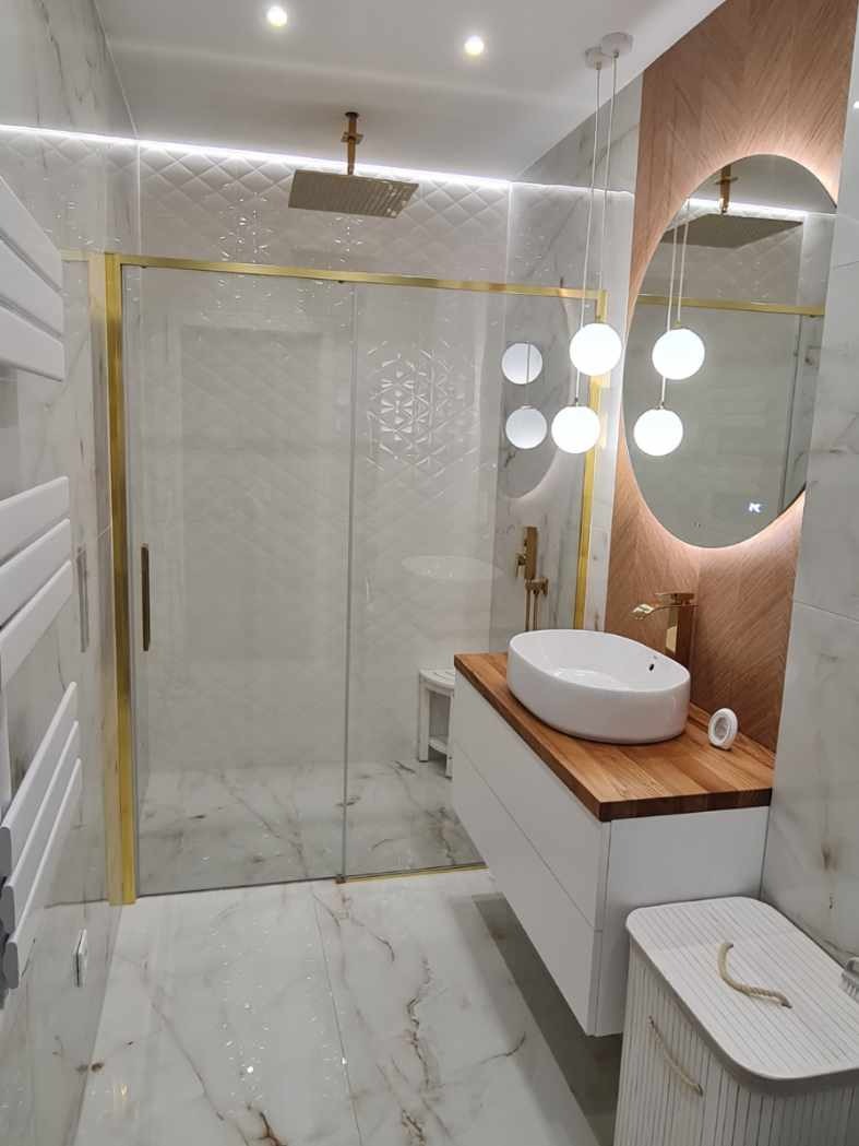 Elegancka łazienka z kabiną prysznicową w złotej ramie, białą umywalką na drewnianym blacie i okrągłym lustrem z podświetleniem. Marmurowe płytki na podłodze i ścianach.