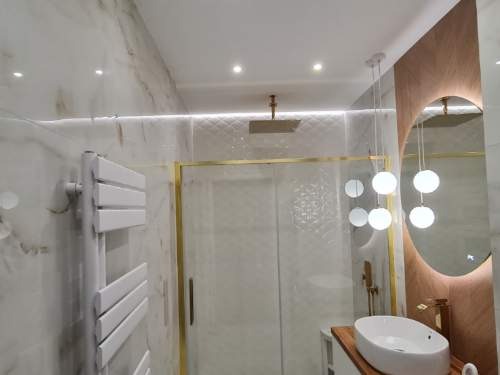 Elegancka łazienka z marmurowymi ścianami, kabiną prysznicową w złotej ramie, owalnym lustrem z podświetleniem i dekoracyjnymi lampami wiszącymi.