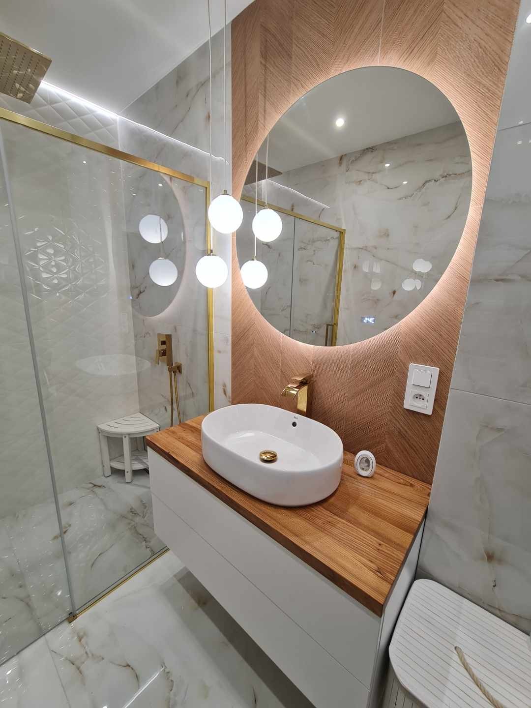 Nowoczesna łazienka z owalnym lustrem podświetlonym LED, białą umywalką na drewnianym blacie i kabiną prysznicową ze złotymi akcentami.
