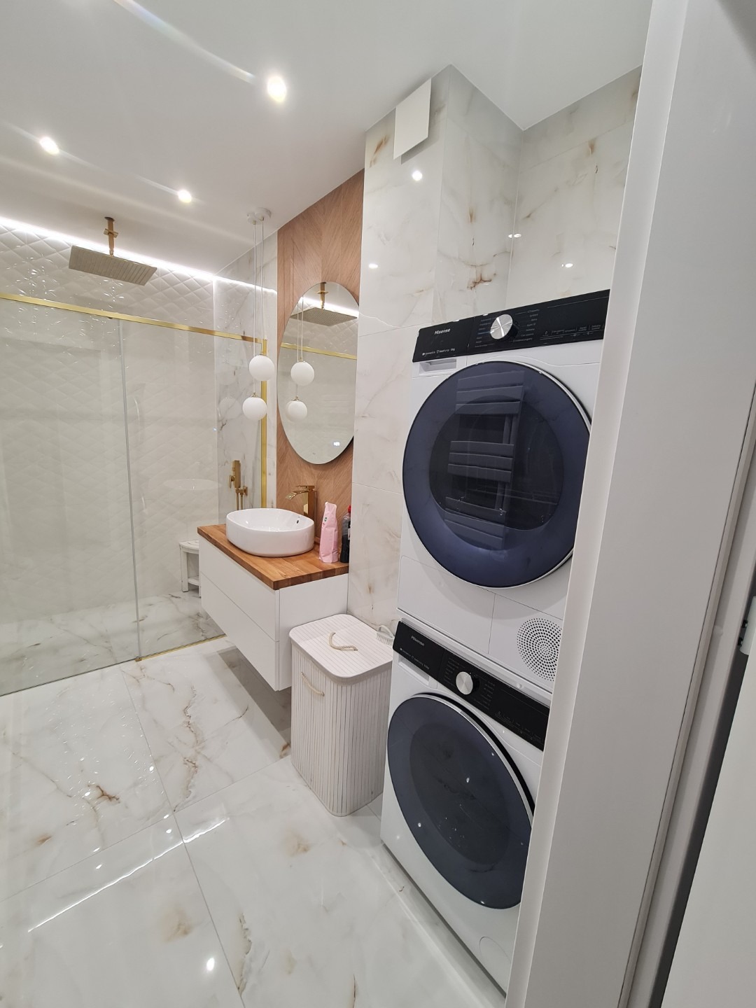Elegancka łazienka z marmurowymi płytkami, wiszącą szafką z drewnianym blatem, okrągłym lustrem i pralko-suszarką w kolumnie. Nowoczesny design.