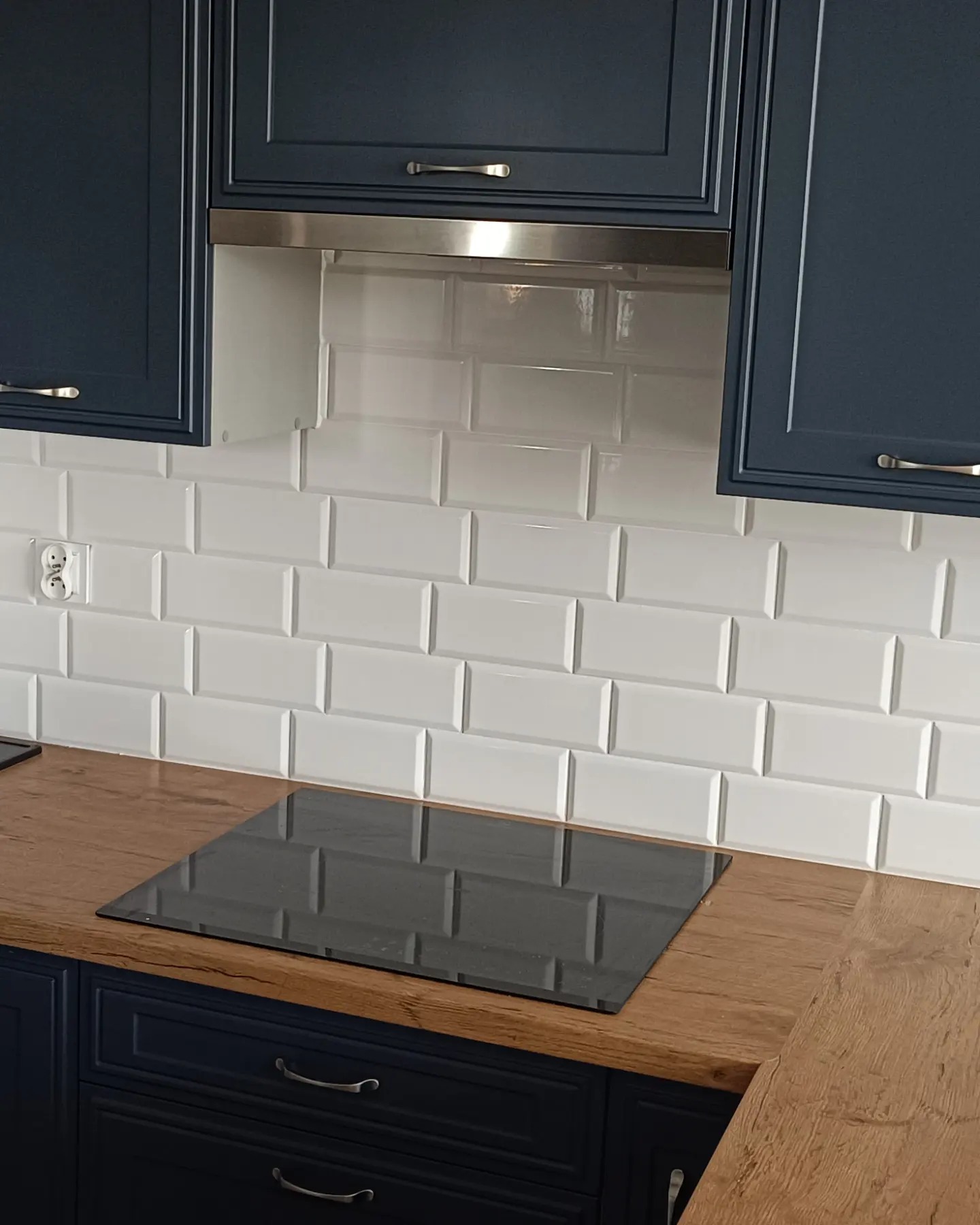 Białe, prostokątne płytki na ścianie nad blatem i płytą indukcyjną. Granatowe szafki z uchwytami. Metalowy okap. Gniazdko elektryczne po lewej.