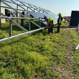 S&M-PV SPÓŁKA Z OGRANICZONĄ ODPOWIEDZIALNOŚCIĄ - Montaż paneli słonecznych na metalowej konstrukcji przez czterech pracowników w odblaskowych kamizelkach i kaskach, na trawiastym terenie w słoneczny dzień.