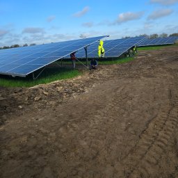 S&M-PV SPÓŁKA Z OGRANICZONĄ ODPOWIEDZIALNOŚCIĄ - Rząd paneli słonecznych na polu z widocznymi śladami opon, pracownicy w odblaskowych kamizelkach przy panelach, błękitne niebo z chmurami w tle.