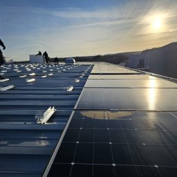 S&M-PV SPÓŁKA Z OGRANICZONĄ ODPOWIEDZIALNOŚCIĄ - Montaż paneli fotowoltaicznych na dachu przemysłowym w słoneczny dzień. Widoczne panele, mocowania i sylwetki instalatorów.