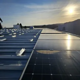 Montaż paneli fotowoltaicznych na dachu przemysłowym w słoneczny dzień. Widoczne panele, mocowania i sylwetki instalatorów.