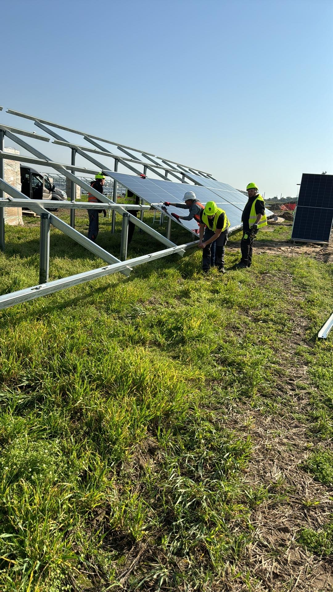 Montaż paneli słonecznych na metalowej konstrukcji przez czterech pracowników w odblaskowych kamizelkach i kaskach, na trawiastym terenie w słoneczny dzień.