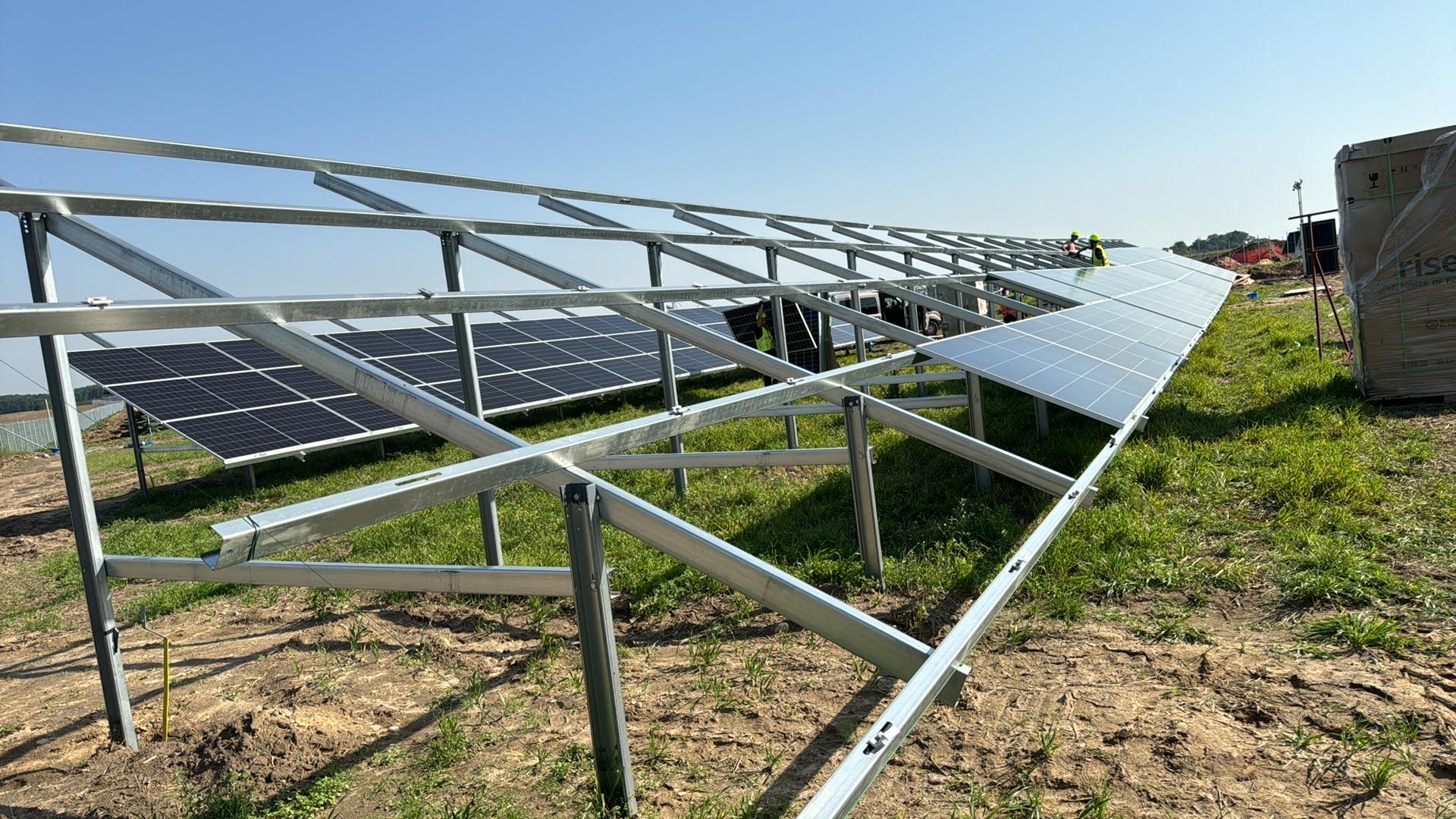 Montaż paneli słonecznych na metalowej konstrukcji na polu; widoczni pracownicy w żółtych kaskach.