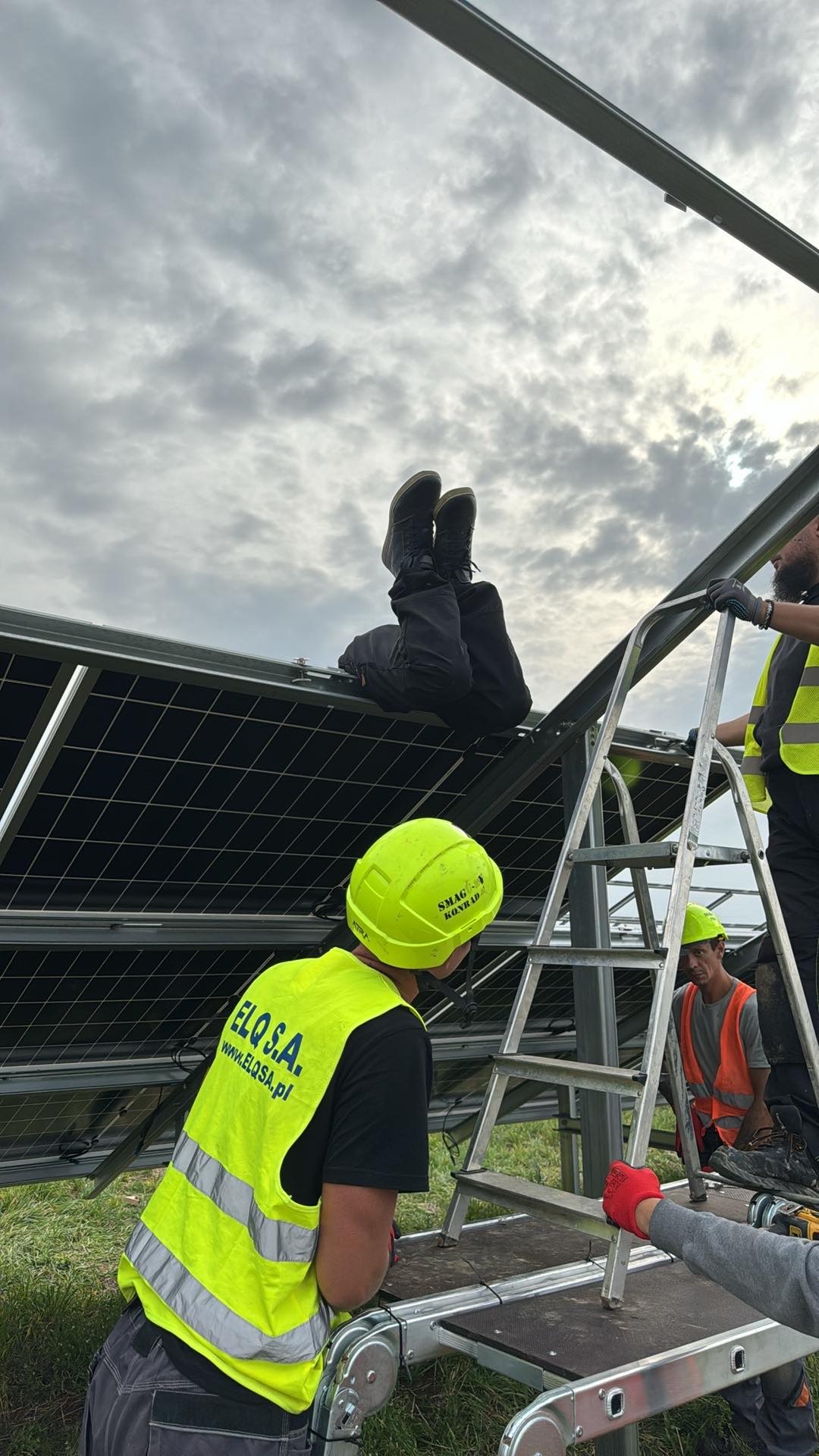 Montaż paneli słonecznych: pracownik w czarnych butach siedzi na panelach, drugi montuje, widoczna drabina i pochmurne niebo.