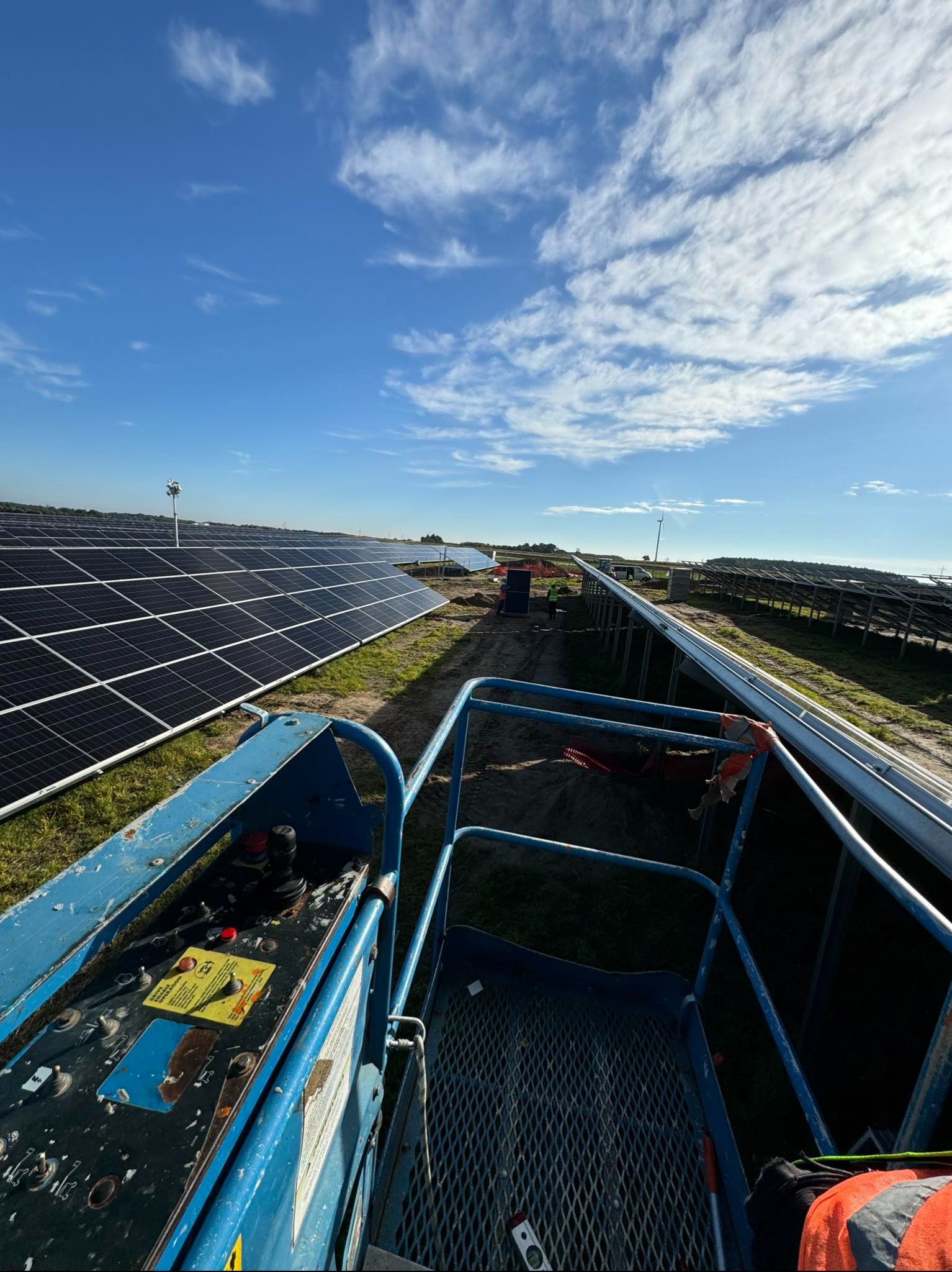 Widok z podnośnika koszowego na rozległą farmę paneli słonecznych pod błękitnym niebem z chmurami, widoczne szyny montażowe w trakcie instalacji.