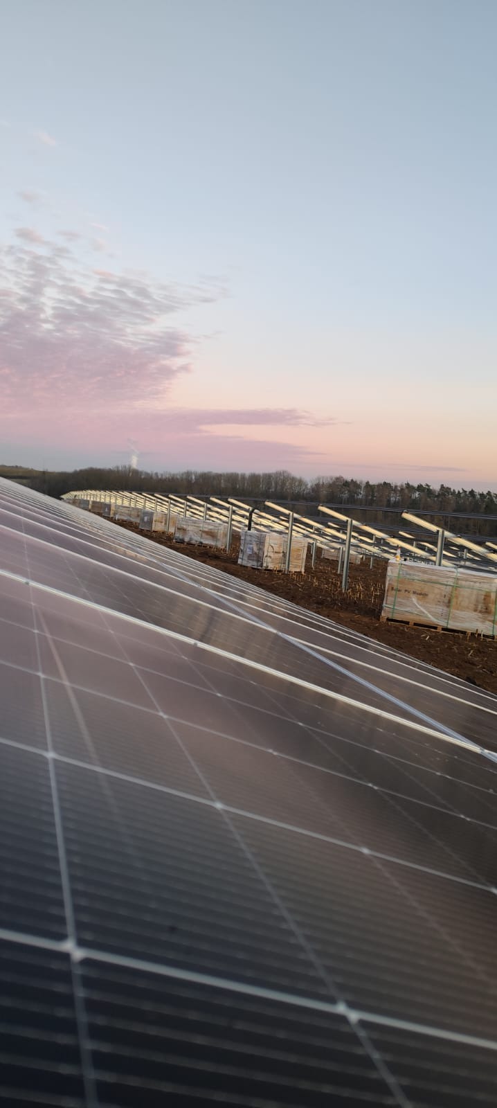 Rząd paneli słonecznych w trakcie montażu o zmierzchu, widoczne stalowe konstrukcje wsporcze i palety z modułami w tle.