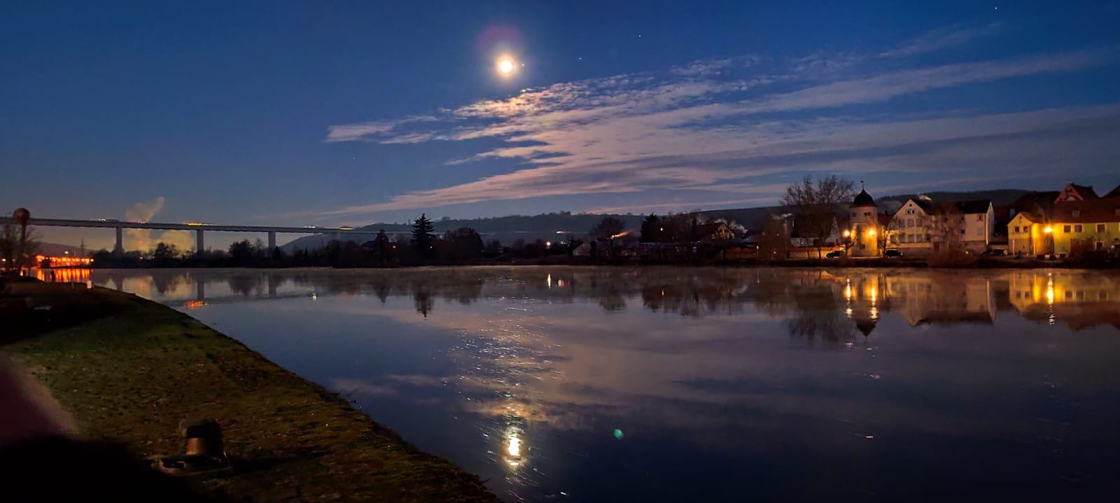 Nocny krajobraz: rzeka, most z oświetleniem LED, zabudowania i księżyc w chmurach, odbicie w wodzie.