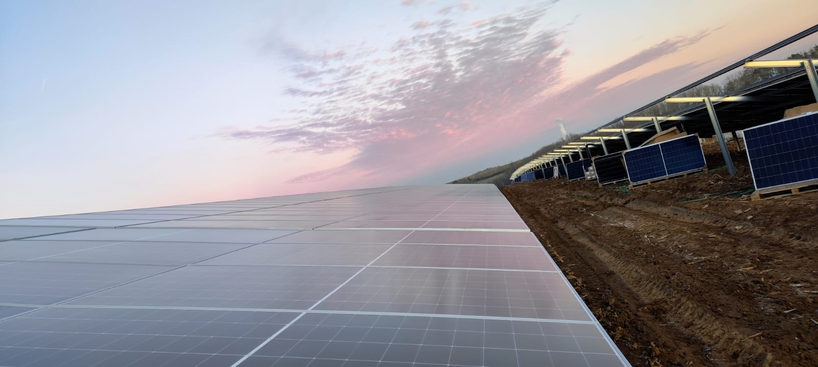 Rozległa farma paneli słonecznych w trakcie montażu, widok z dołu pod kątem, ziemia z koleinami, zachodzące słońce z chmurami w tle.