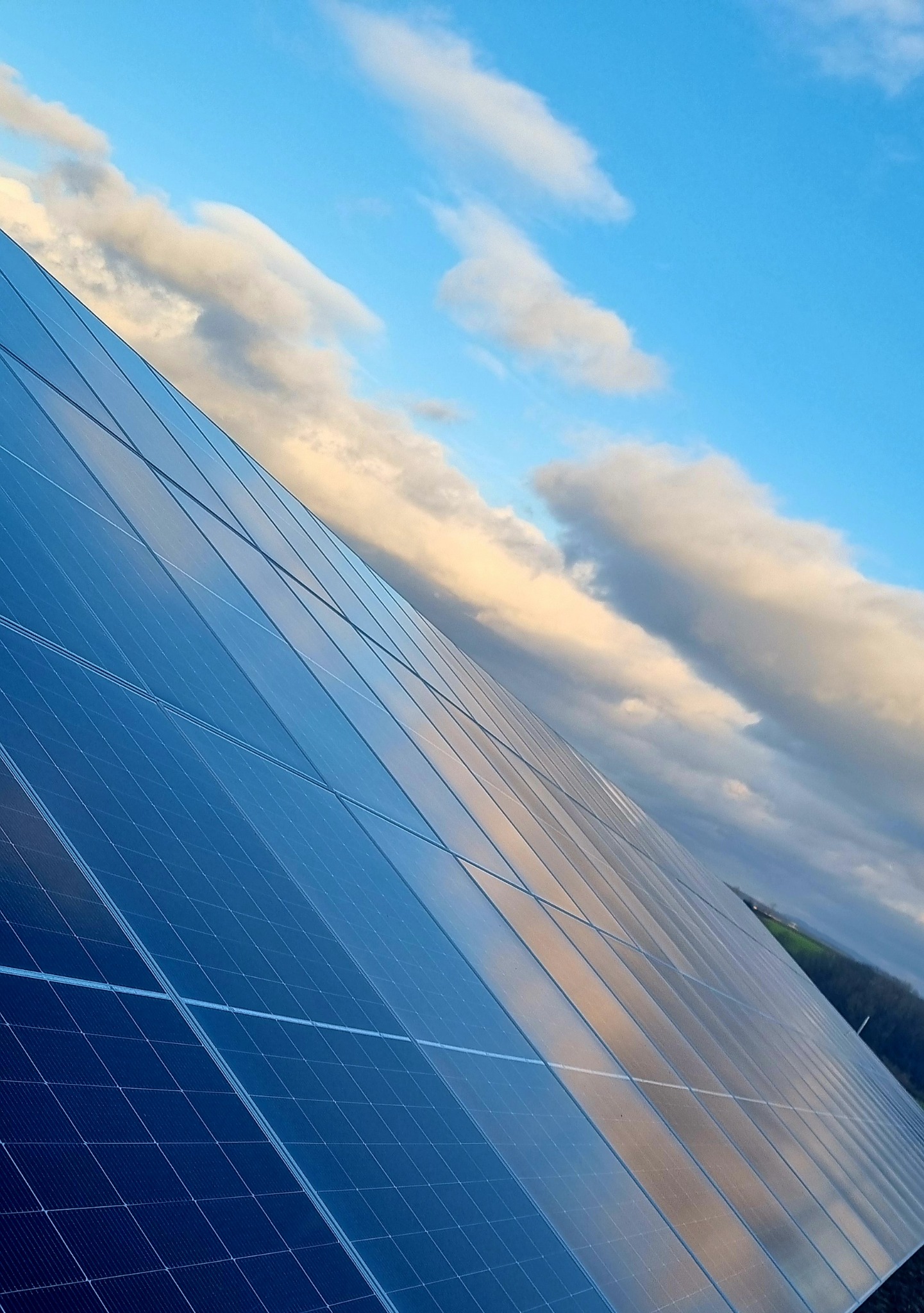 Panel słoneczny odbijający błękitne niebo z białymi chmurami, widok pod kątem, zieleń w tle.