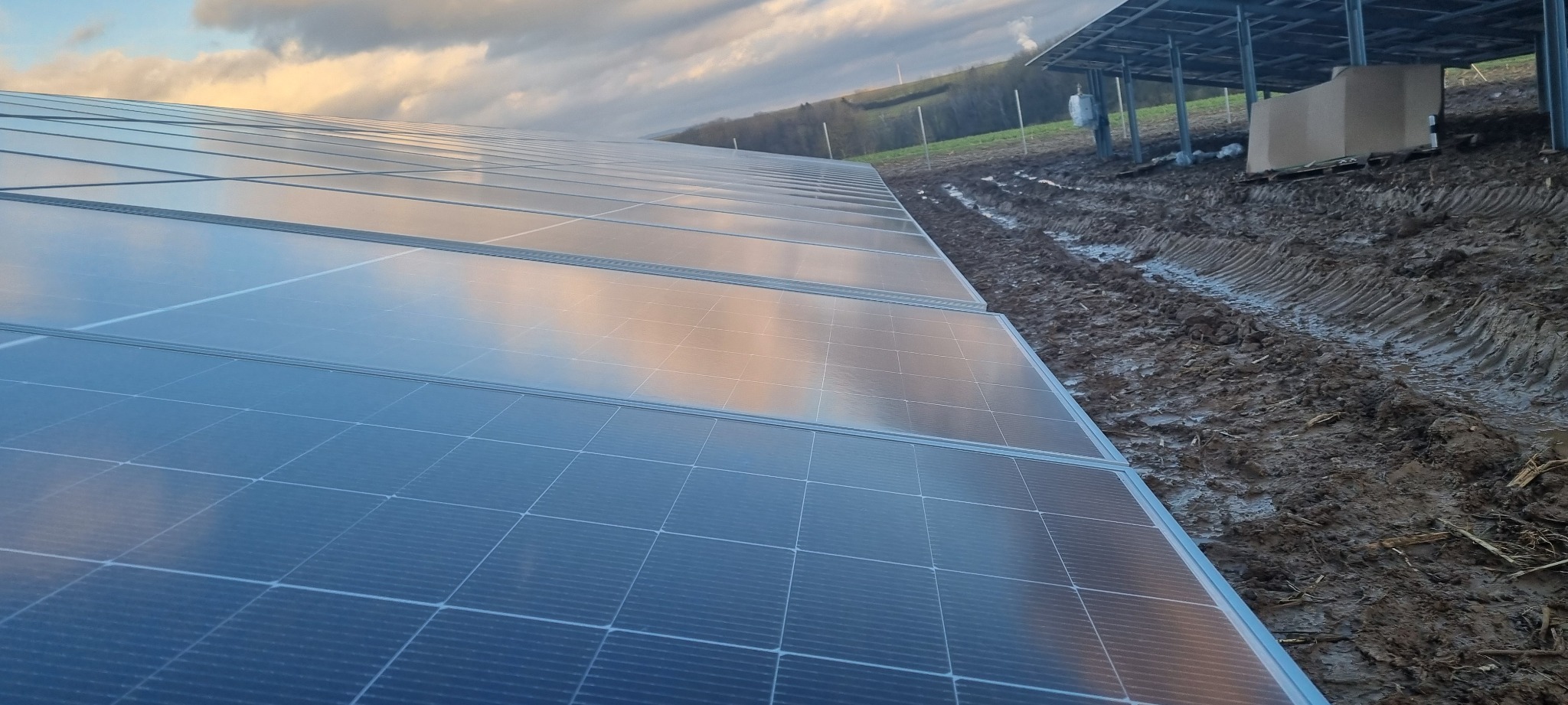 Panel słoneczny w trakcie instalacji, z odbiciem chmur na powierzchni, obok błotnistej ziemi i konstrukcji wsporczej.