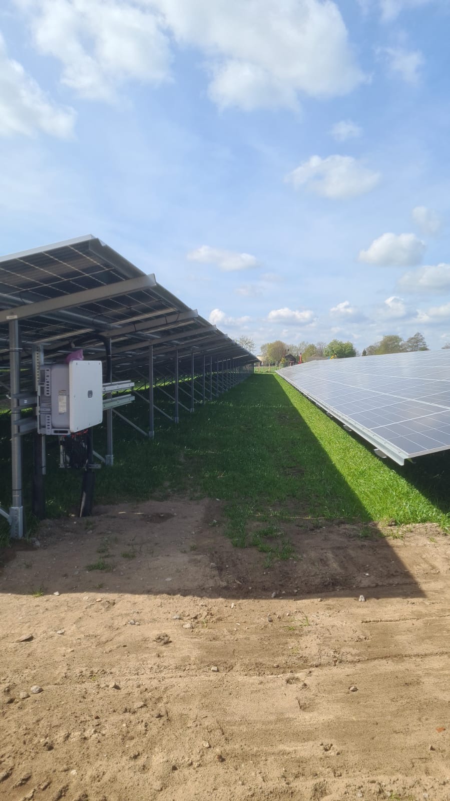 Rząd paneli słonecznych zamontowanych na metalowej konstrukcji na trawiastym terenie, z widocznym inwerterem i niebem z chmurami w tle.