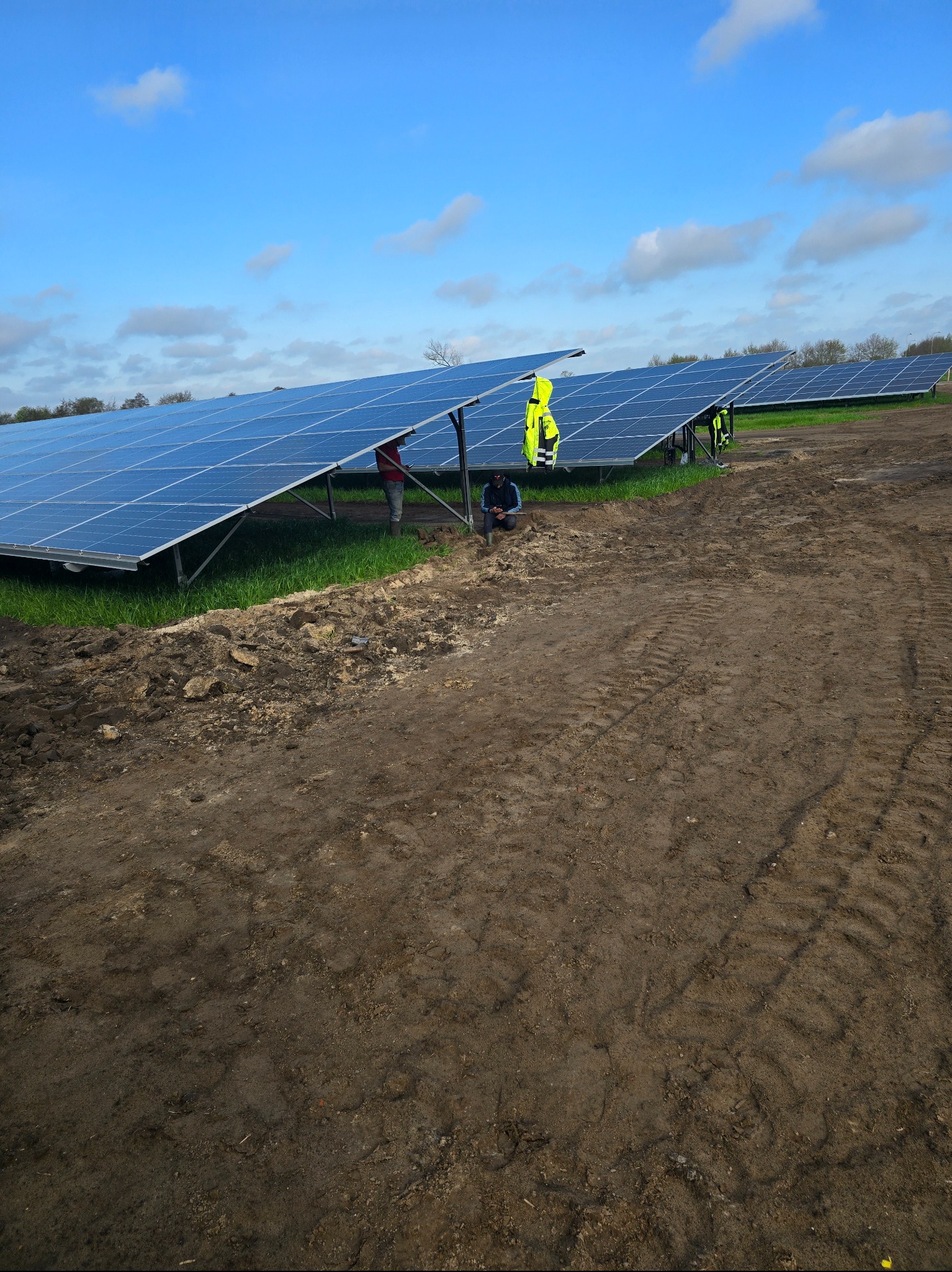 Rząd paneli słonecznych na polu z widocznymi śladami opon, pracownicy w odblaskowych kamizelkach przy panelach, błękitne niebo z chmurami w tle.