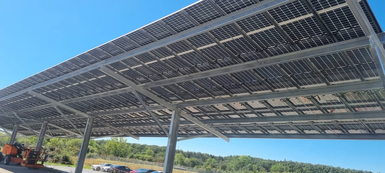 Instalacja paneli fotowoltaicznych na konstrukcji wsporczej, widok od dołu. Na dole widać zaparkowane samochody i zieleń. Błękitne niebo w tle.