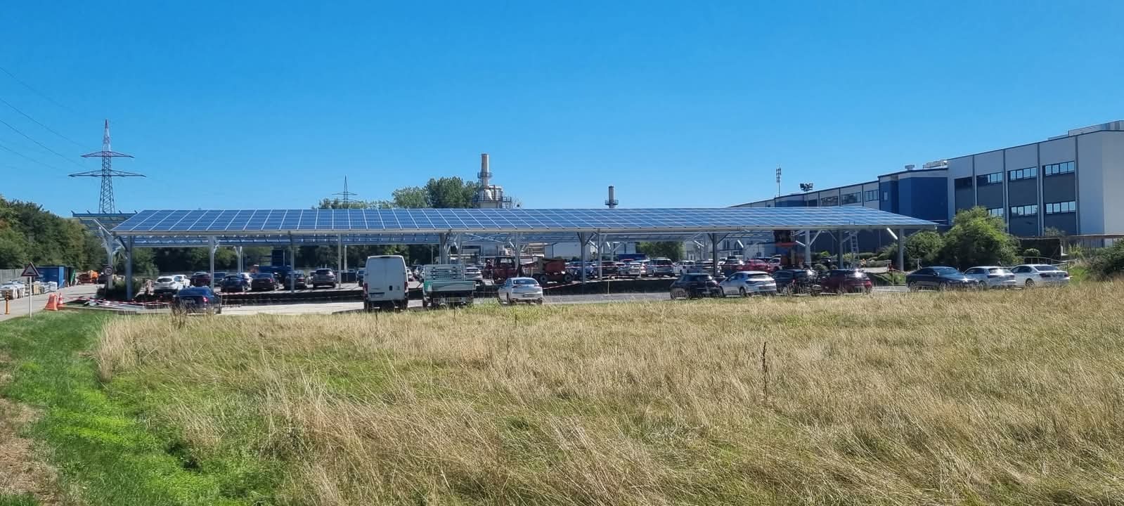 Parking z zadaszeniem z paneli fotowoltaicznych w słoneczny dzień. W tle budynek przemysłowy i słup wysokiego napięcia.