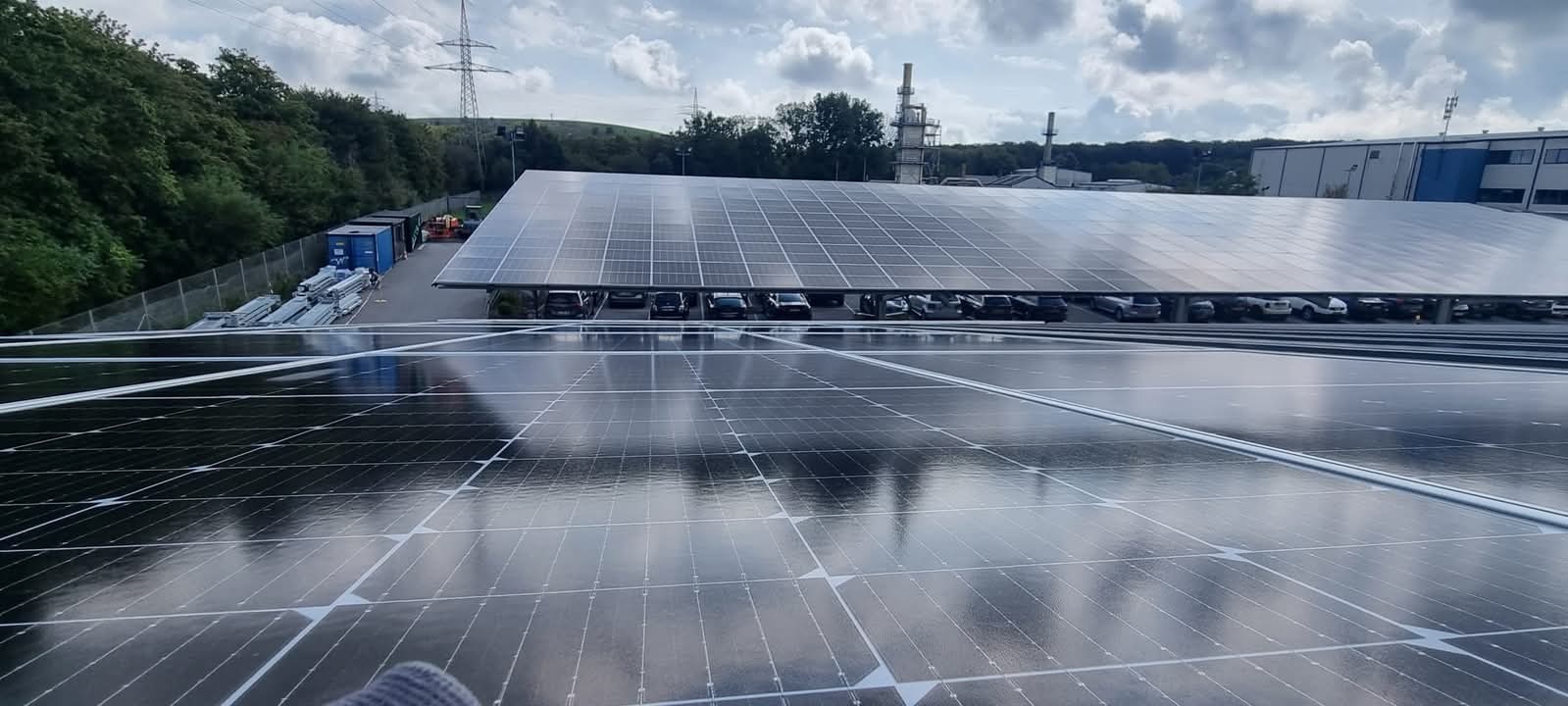 Rozległa instalacja fotowoltaiczna na dachu parkingu, odbijająca pochmurne niebo. W tle budynek przemysłowy i słupy wysokiego napięcia.