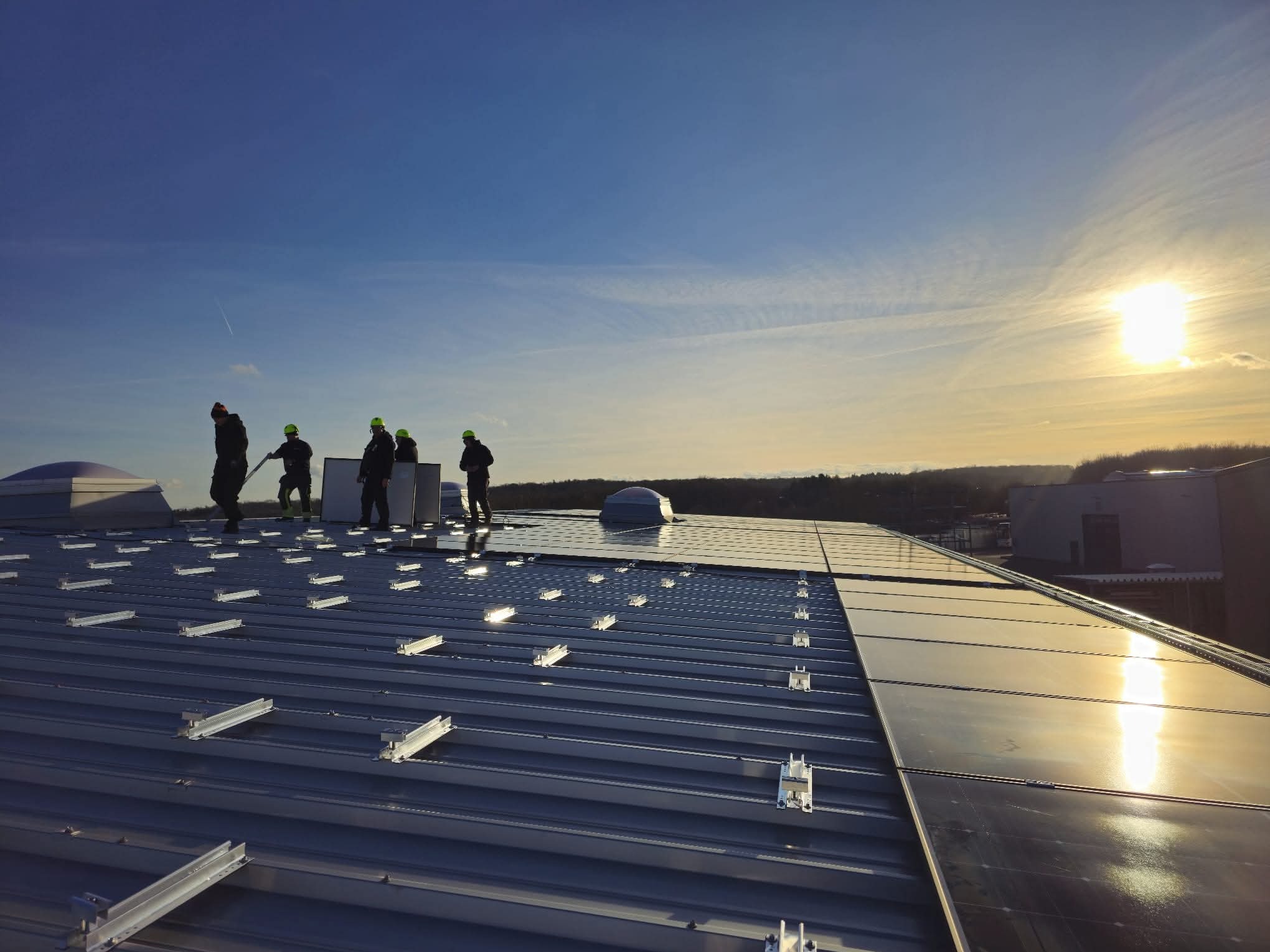 Instalacja paneli słonecznych na dachu przemysłowym; ekipa w kaskach montuje panele; widoczne mocowania i odbicia słońca na panelach.
