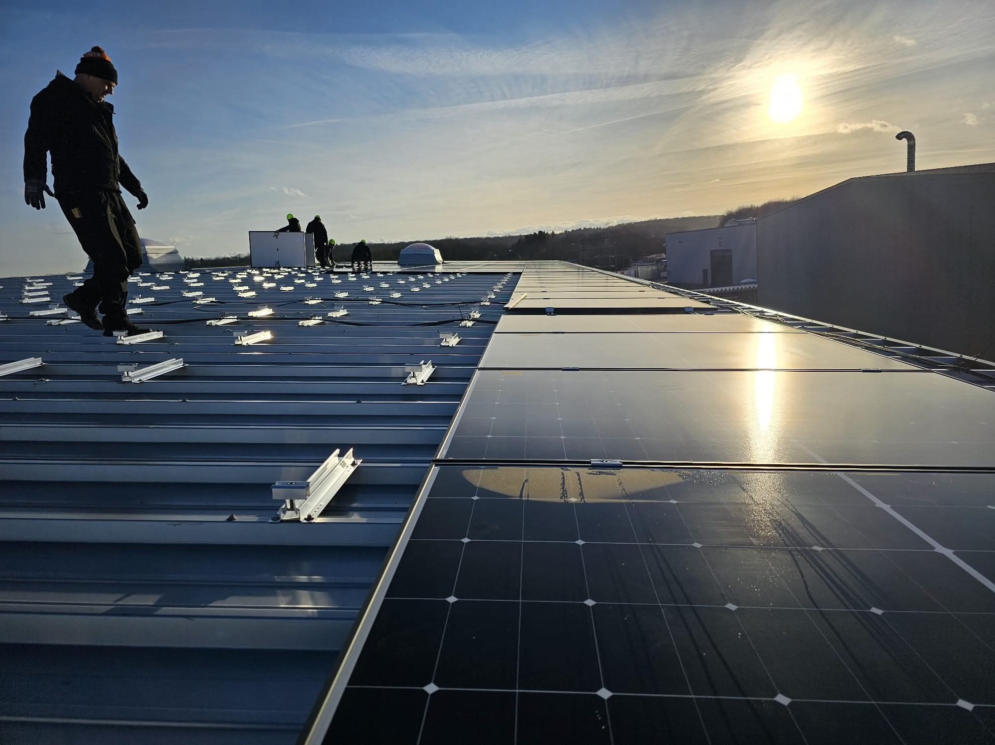 Montaż paneli fotowoltaicznych na dachu przemysłowym w słoneczny dzień. Widoczne panele, mocowania i sylwetki instalatorów.