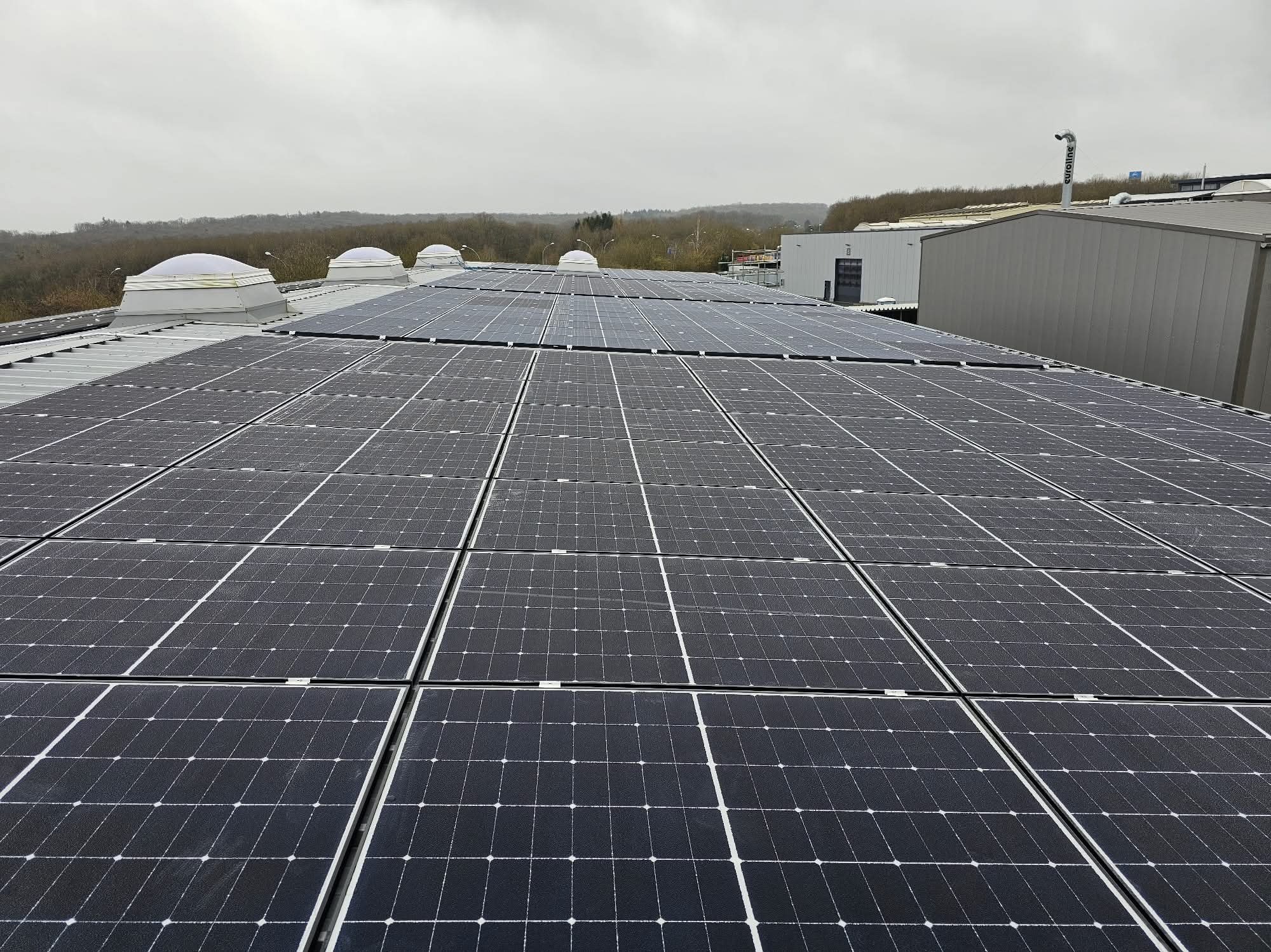 Rozległa instalacja fotowoltaiczna na dachu budynku przemysłowego w pochmurny dzień. Widoczne świetliki dachowe i elementy otoczenia.
